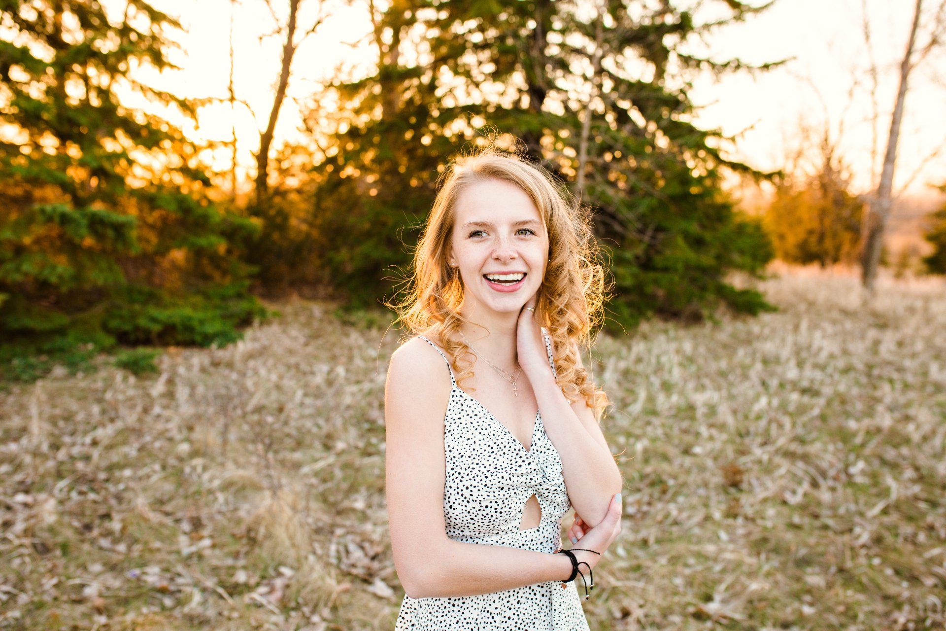 Woman with curly hair smiles, outdoors in front of trees, wearing a white patterned dress.