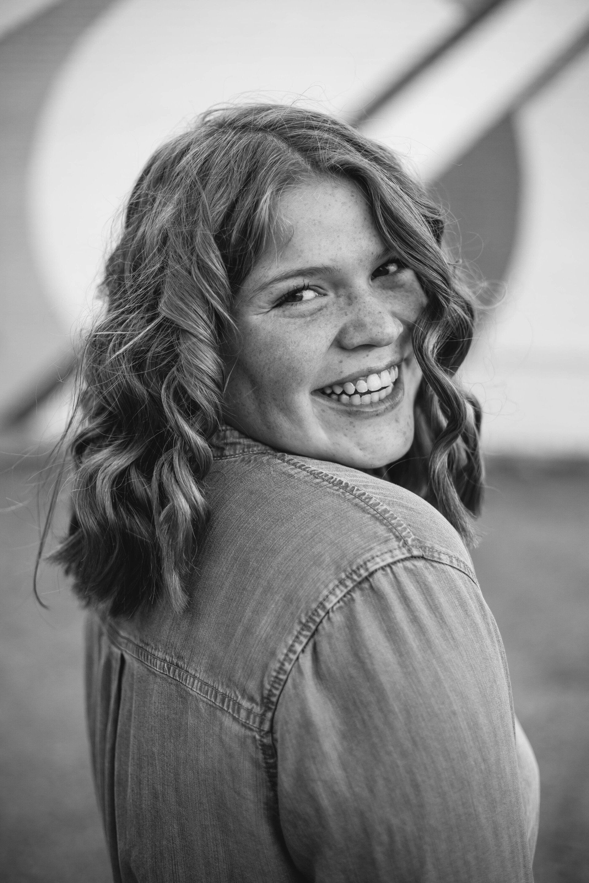 Smiling woman with curly hair, wearing denim shirt, looking over her shoulder.