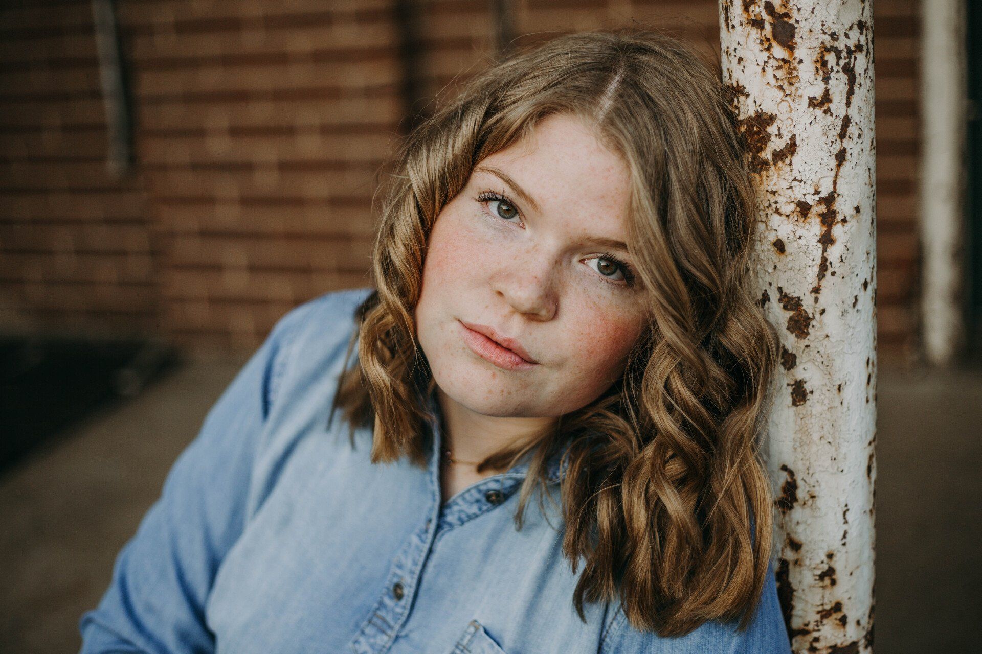 Woman with wavy blonde hair, wearing a blue denim shirt, leaning on a rusty metal pole near a brick wall.