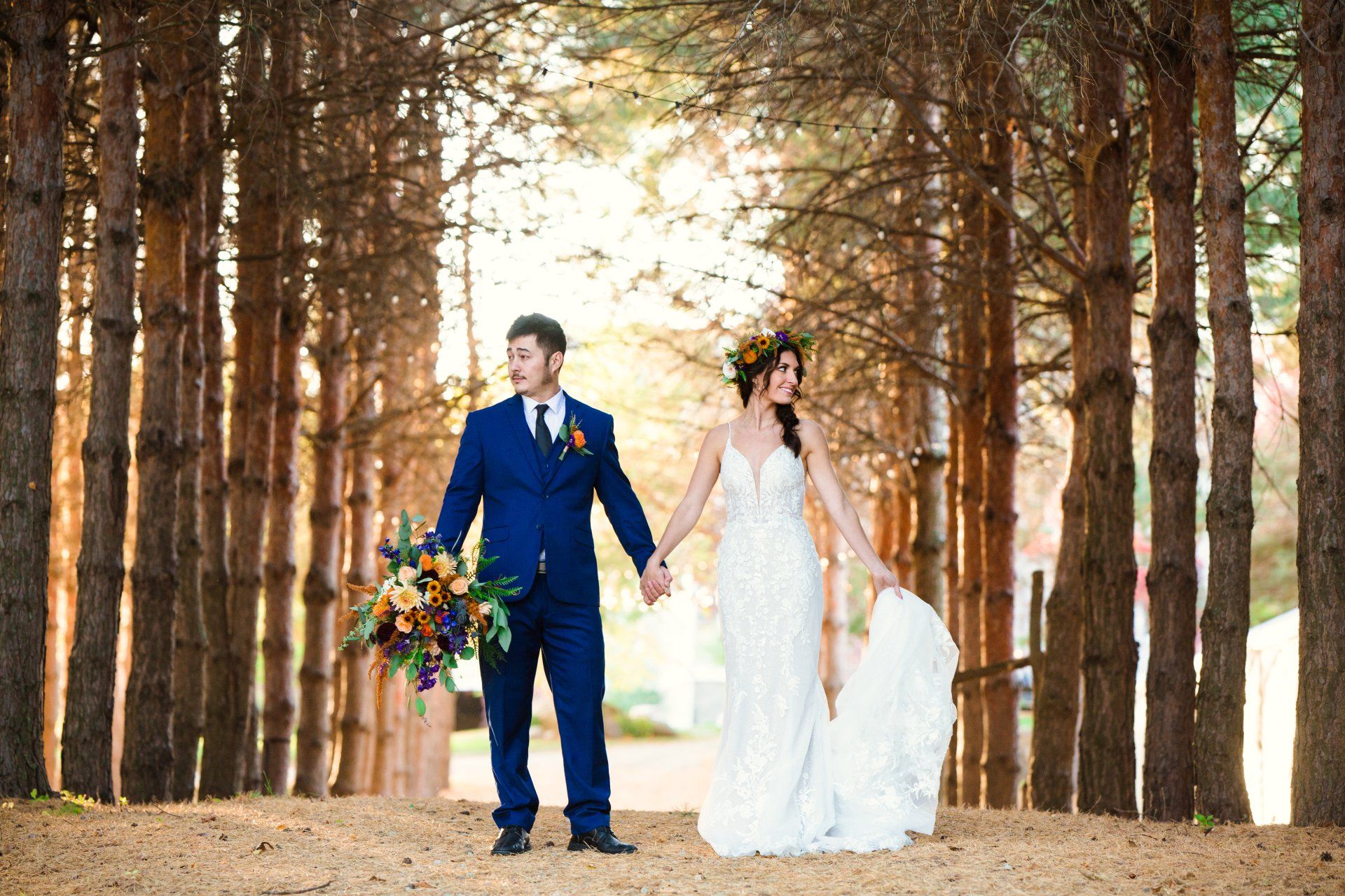 Wedding couple holding hands, standing in a forest path lined with tall trees. Groom in blue suit, bride in white gown.