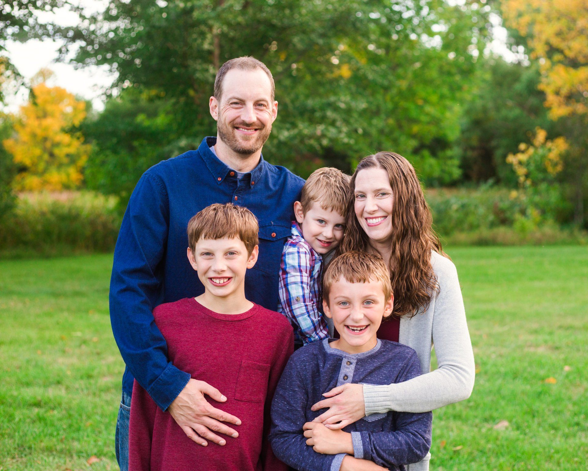 Family of five smiling in a grassy field: two boys hugging, two adults smiling, and a third boy in the middle.