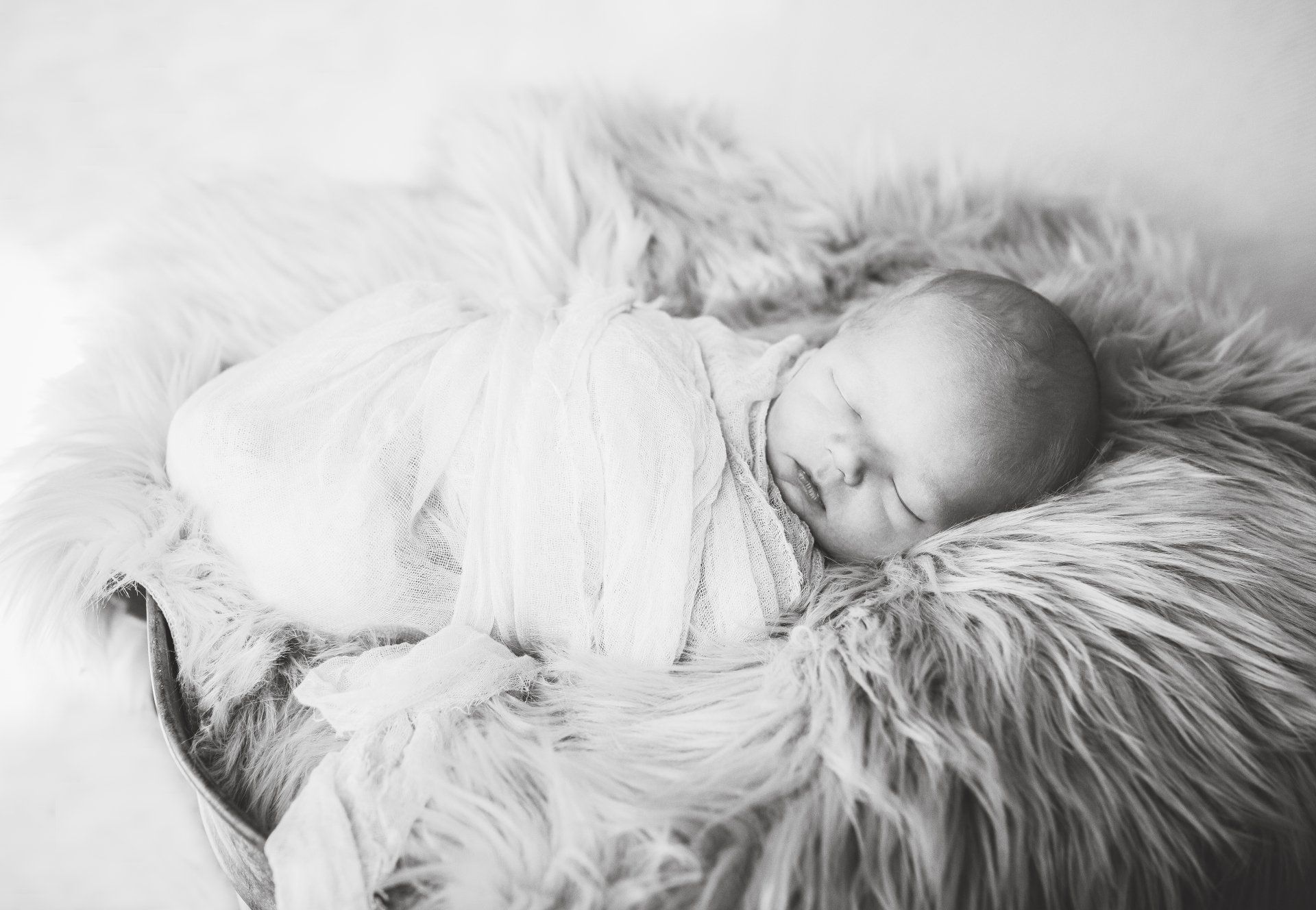 Newborn baby swaddled in white blanket, sleeping on fluffy, light-colored fur.