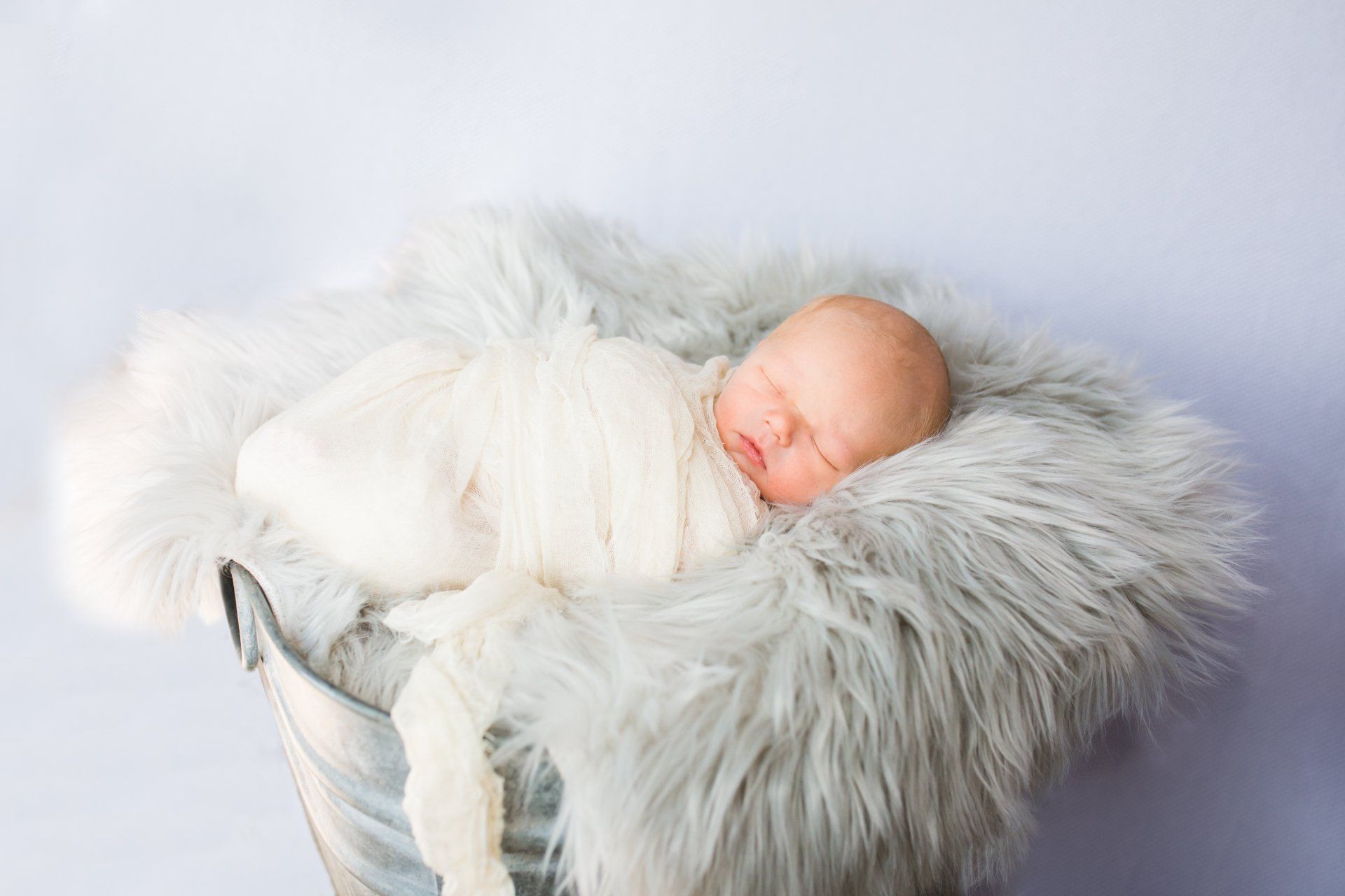 Newborn baby sleeping wrapped in a white blanket, nestled in a gray fur-lined bucket.