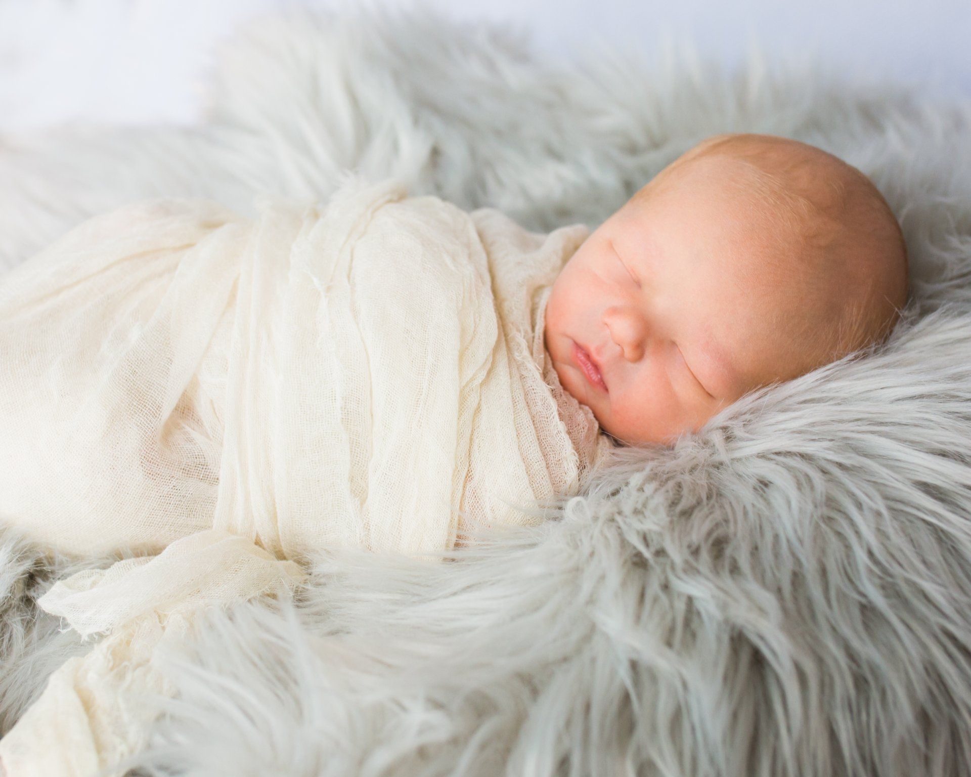 Newborn baby swaddled in cream blanket, resting on soft gray fur.