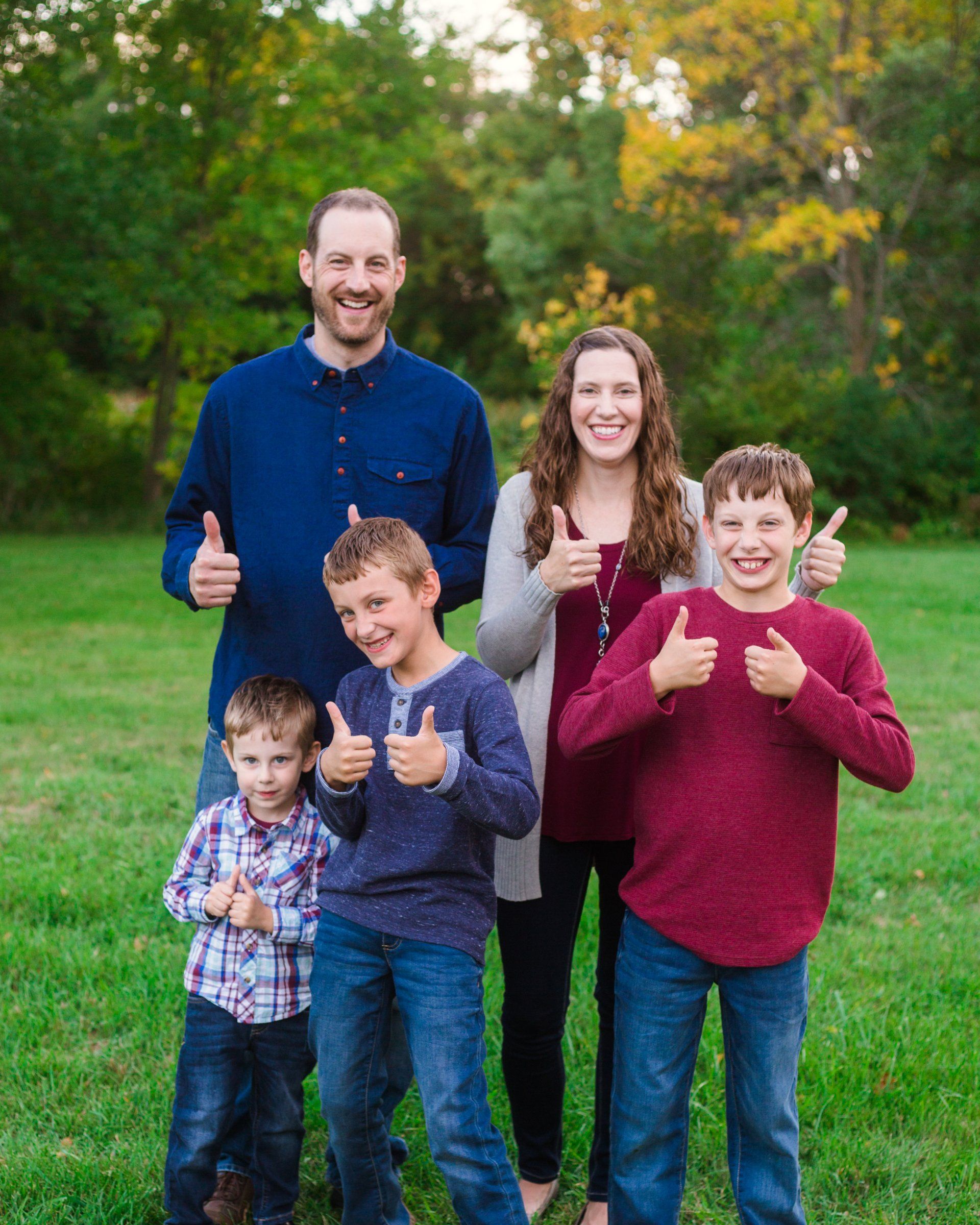 Family of five giving thumbs up in a green grassy area with trees in the background.