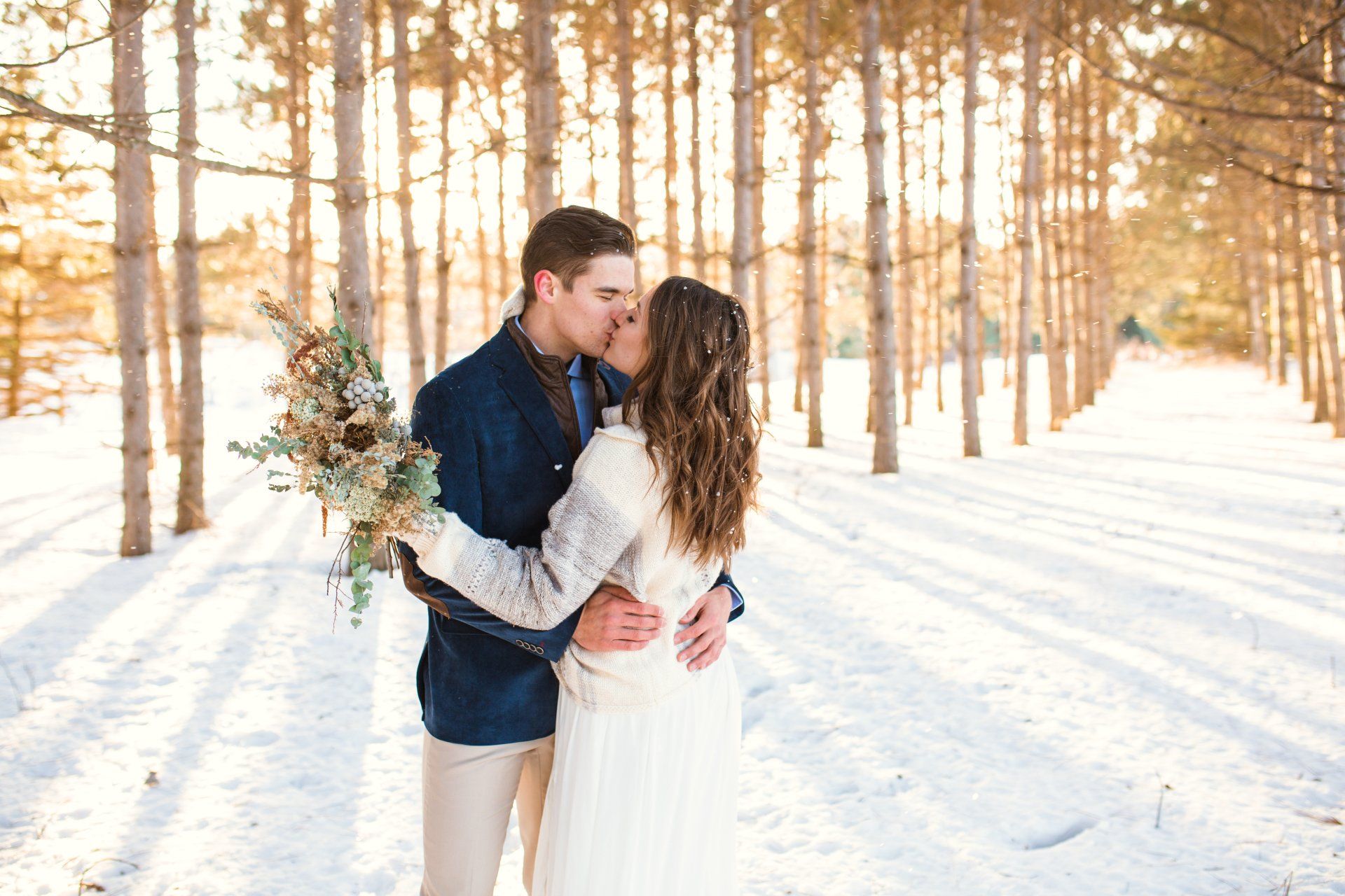 Couple kissing in snow-covered forest. Man in blue coat, woman in wedding dress, bouquet in hand, trees in background.