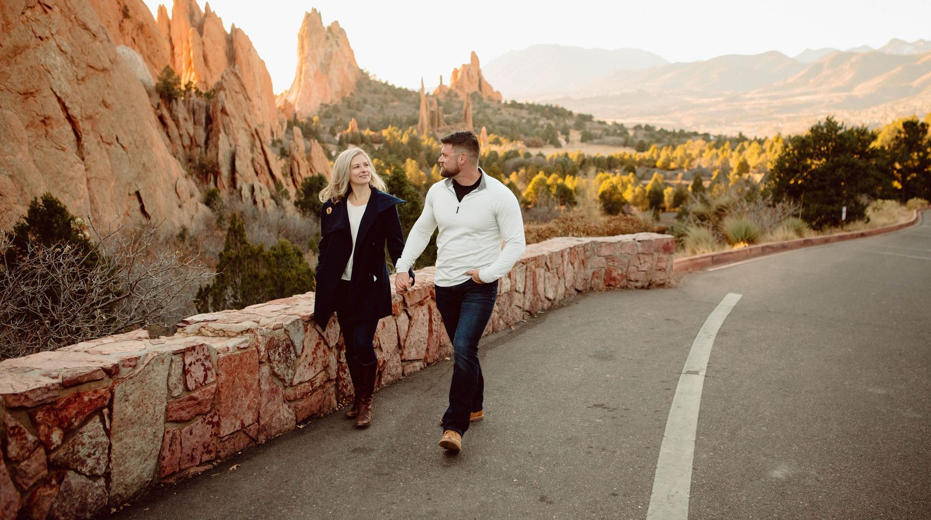 Couple holding hands, walking on a road in a rocky, mountain landscape at sunset.