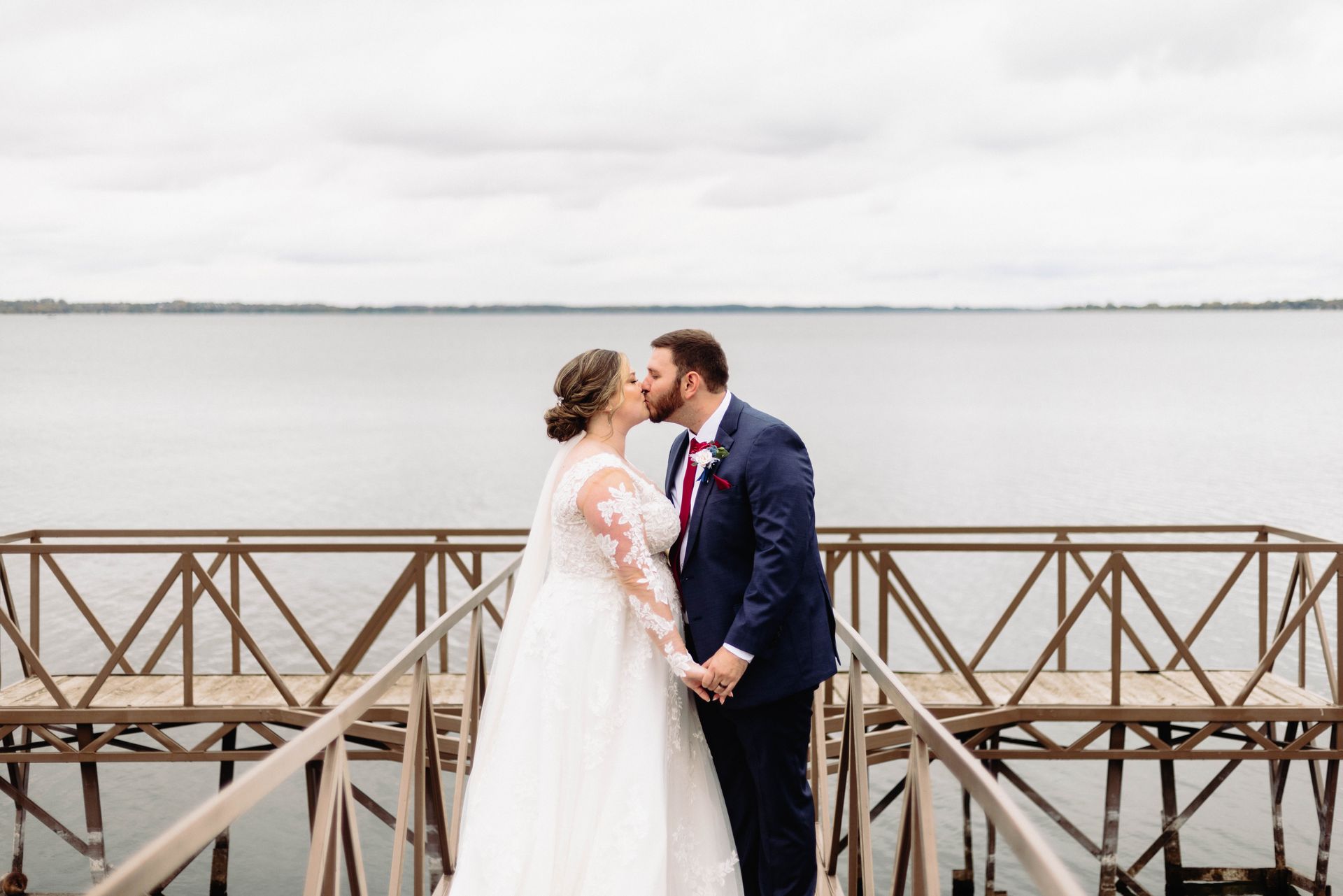 Bride and groom kiss on a wooden pier overlooking water. Cloudy sky, both in wedding attire, holding hands.