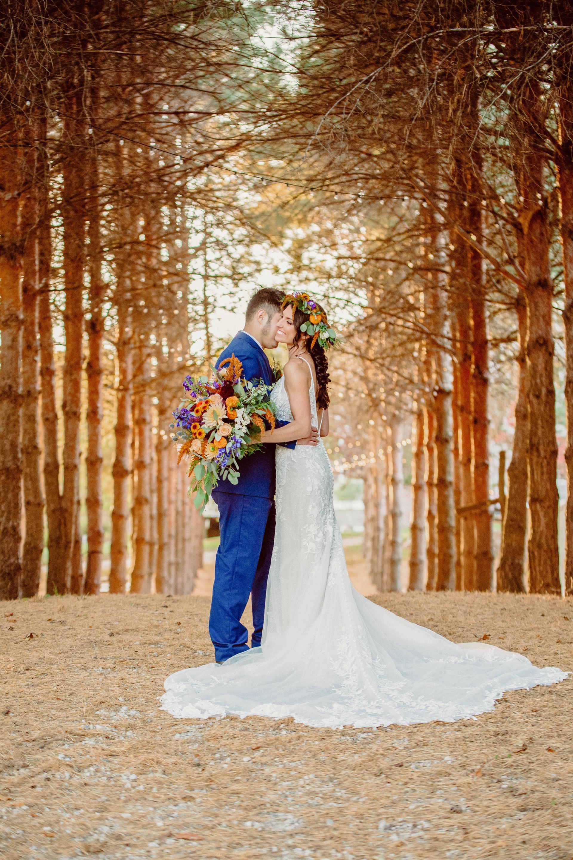 Couple kissing in a forest setting, bride in white dress, groom in blue suit, holding a bouquet.