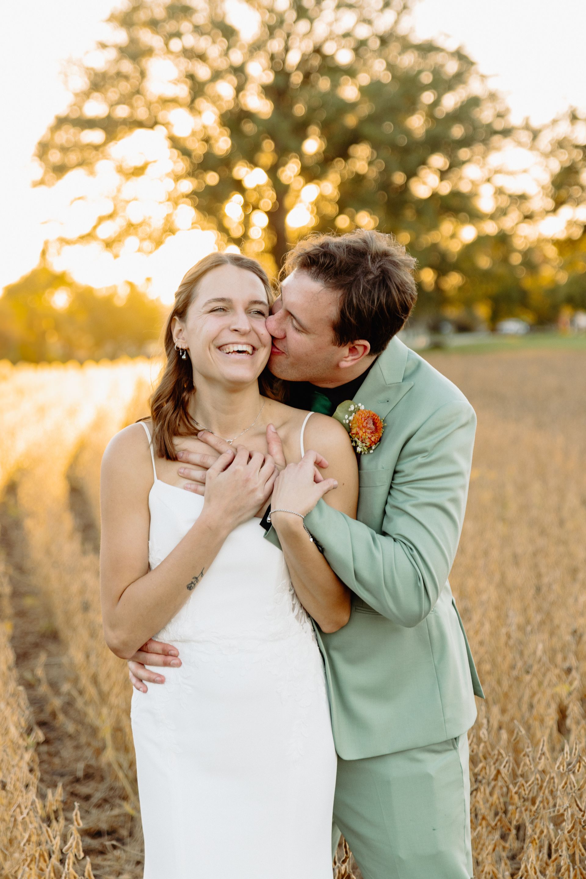 Wedding couple in a field; man kisses woman's cheek, both smiling. Warm light, green suit, white dress.
