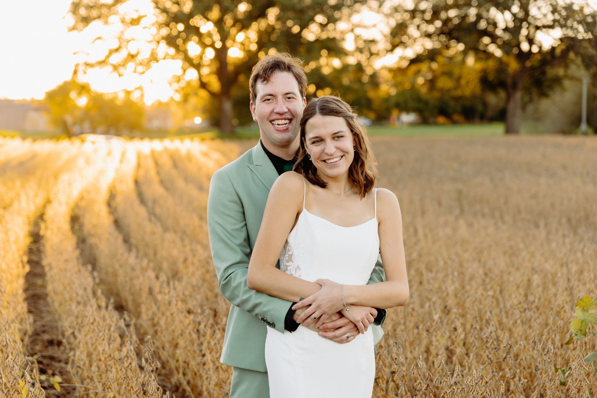 A newlywed couple embraces in a field at sunset. The woman wears a white dress, the man a green suit, both smiling.