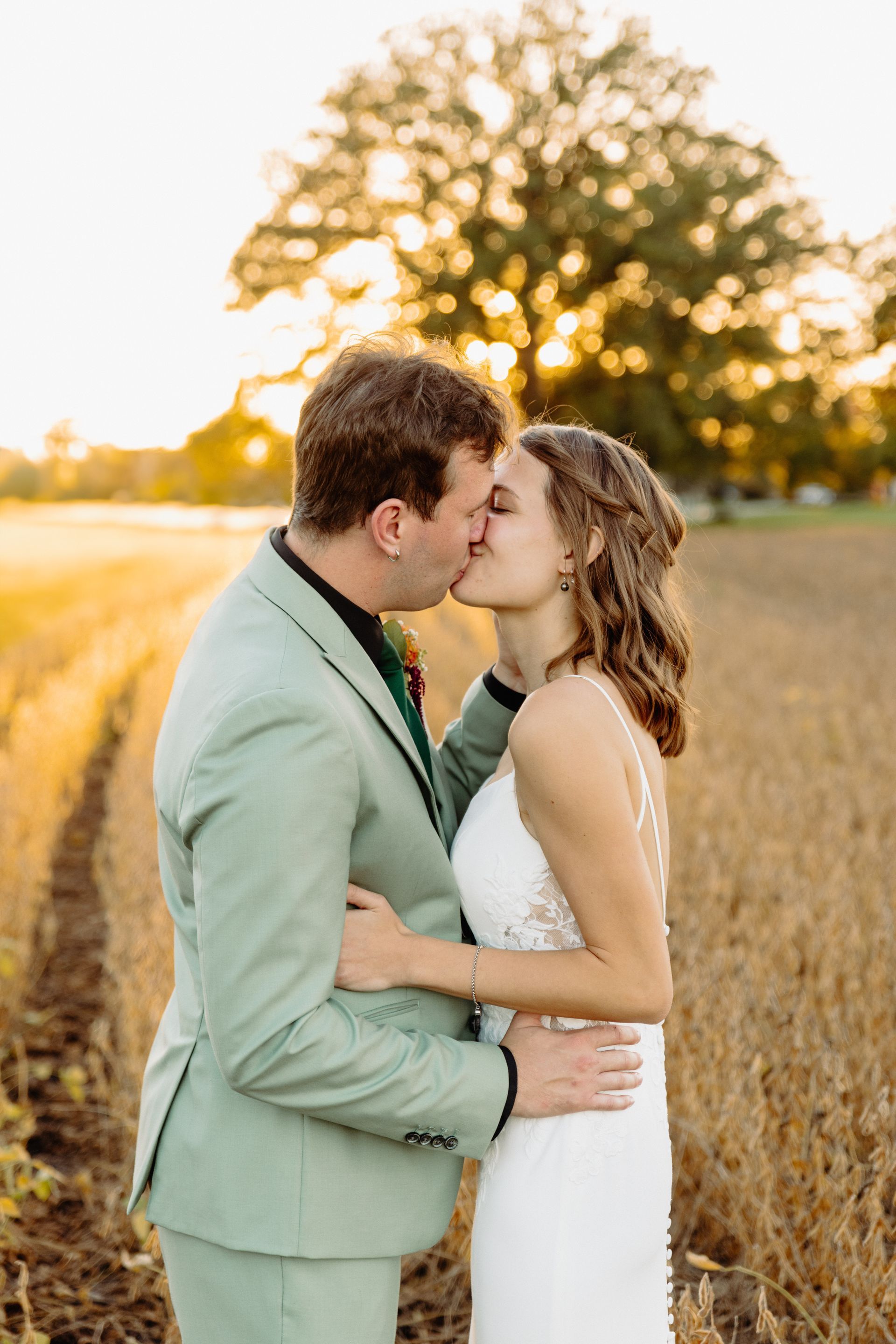 Newlyweds kissing in a field at sunset; the groom wears a green suit, the bride a white dress.