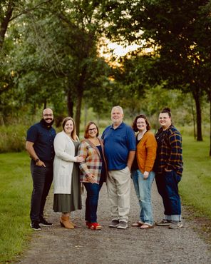 Family of six stands on a path, smiling, in a wooded area. Evening sunlight.
