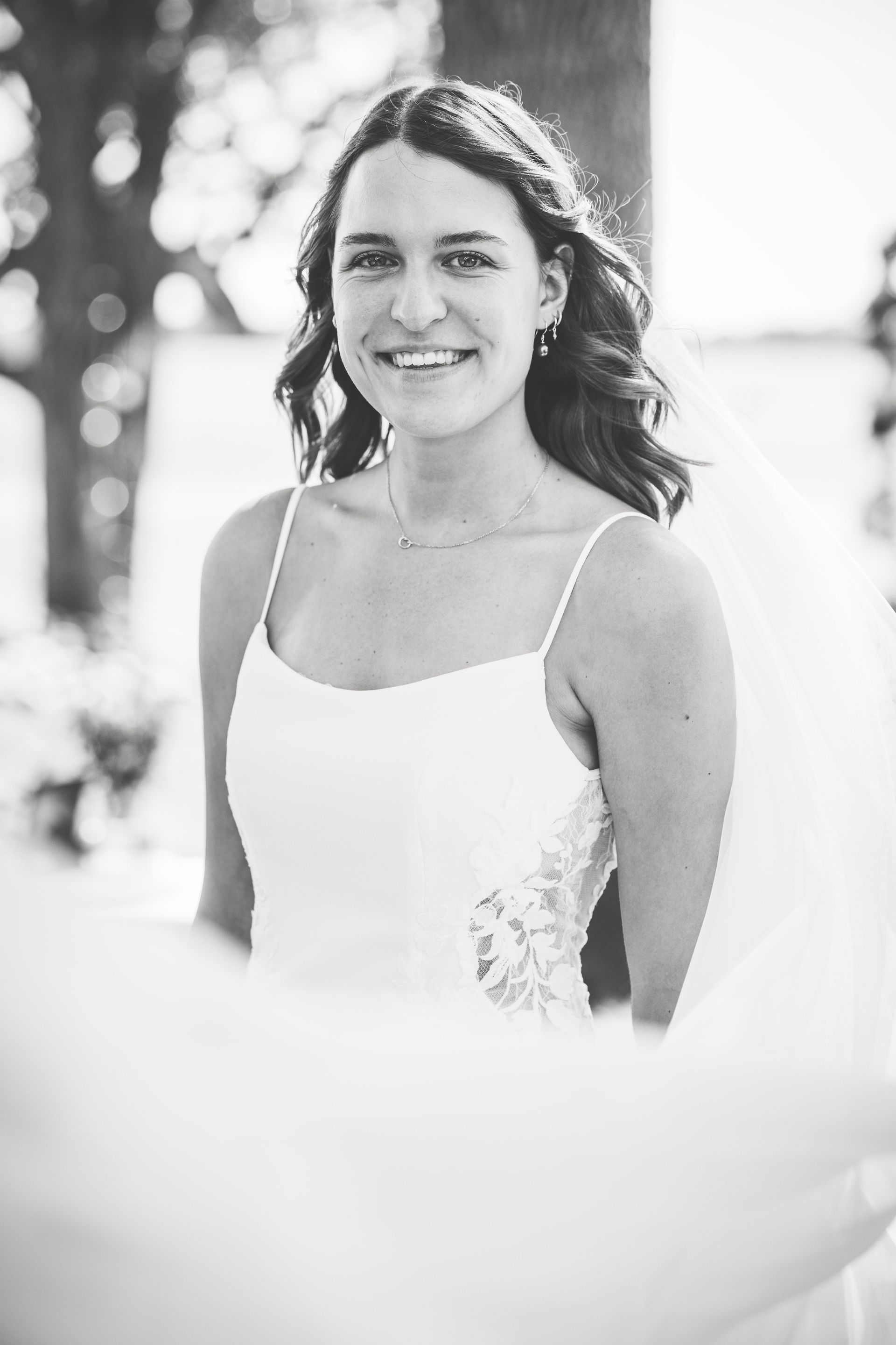 Woman in a white wedding dress smiles outdoors, wearing a veil, looking at the camera.