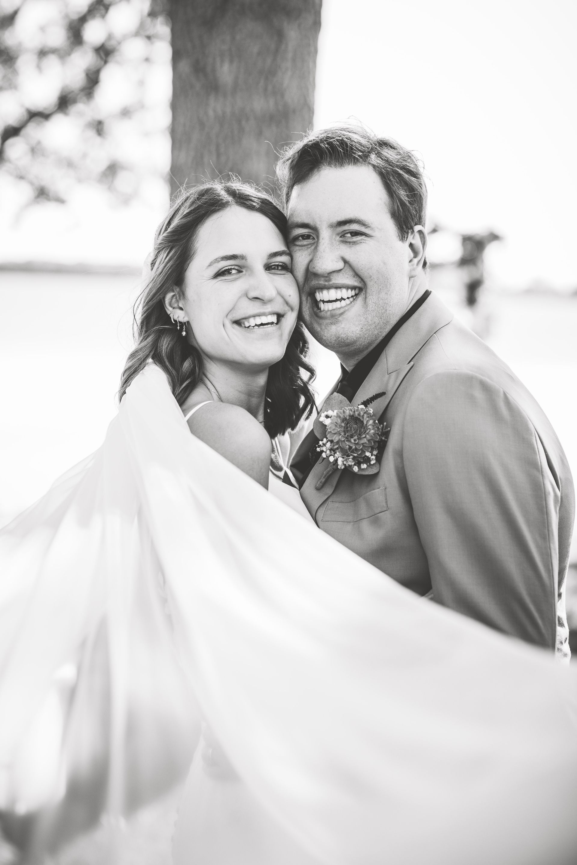 Bride and groom smiling, embracing outdoors; bride's veil billowing; black and white photo.