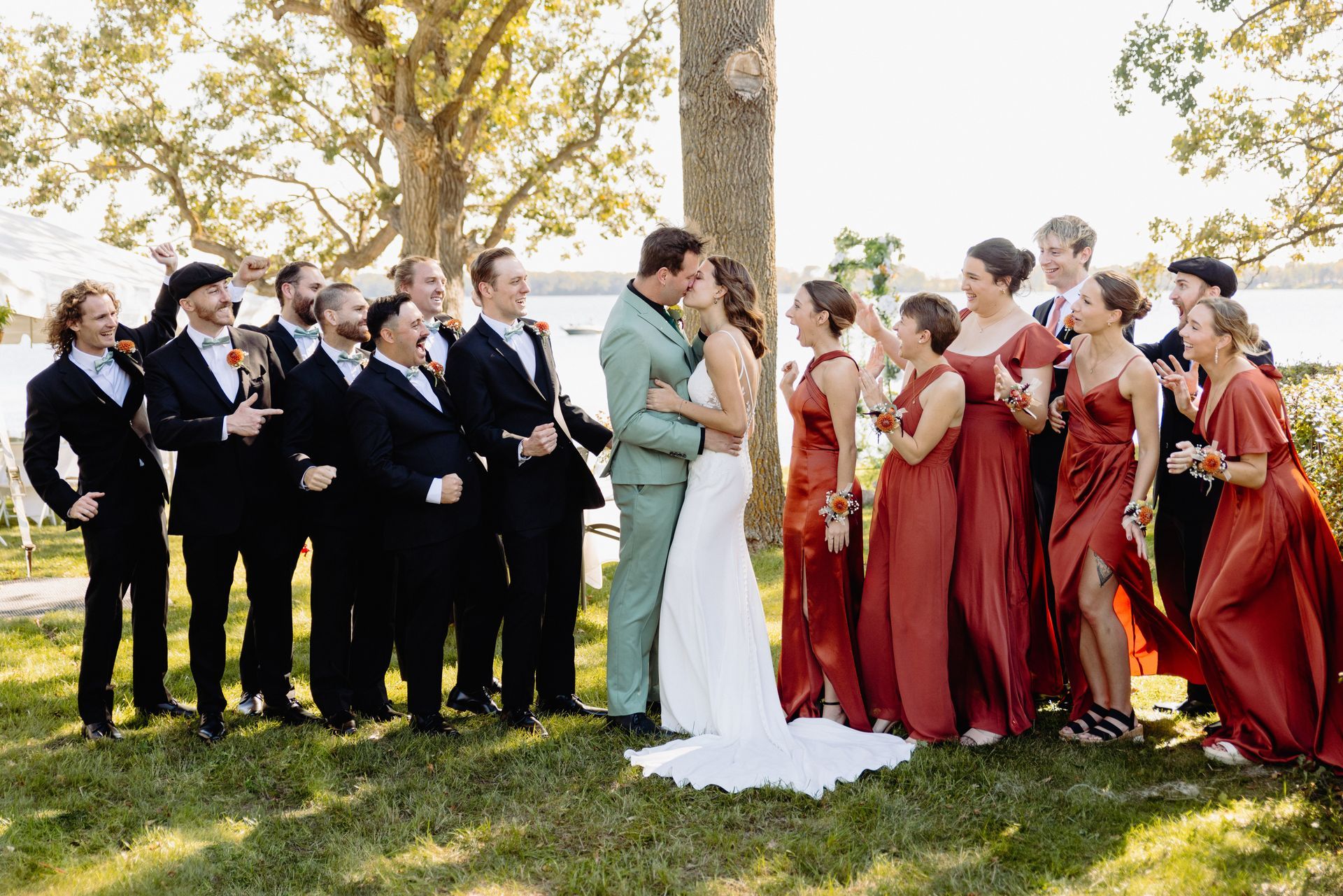 Wedding party: Bride and groom kissing with bridesmaids in red dresses and groomsmen in black suits outdoors.