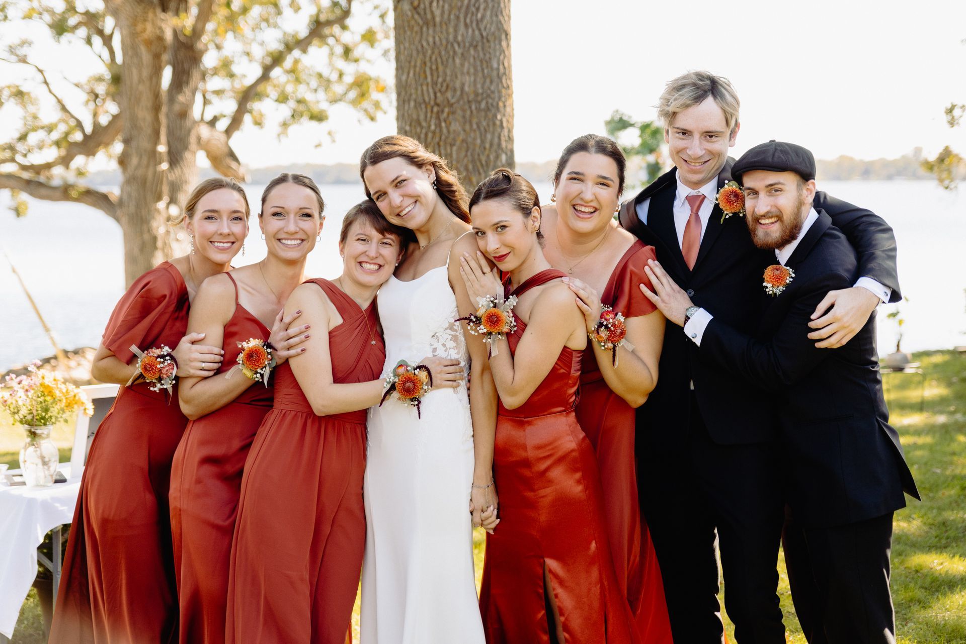 Wedding group: Bride in white dress with bridesmaids in red dresses and groomsmen in suits, posed outside near water.
