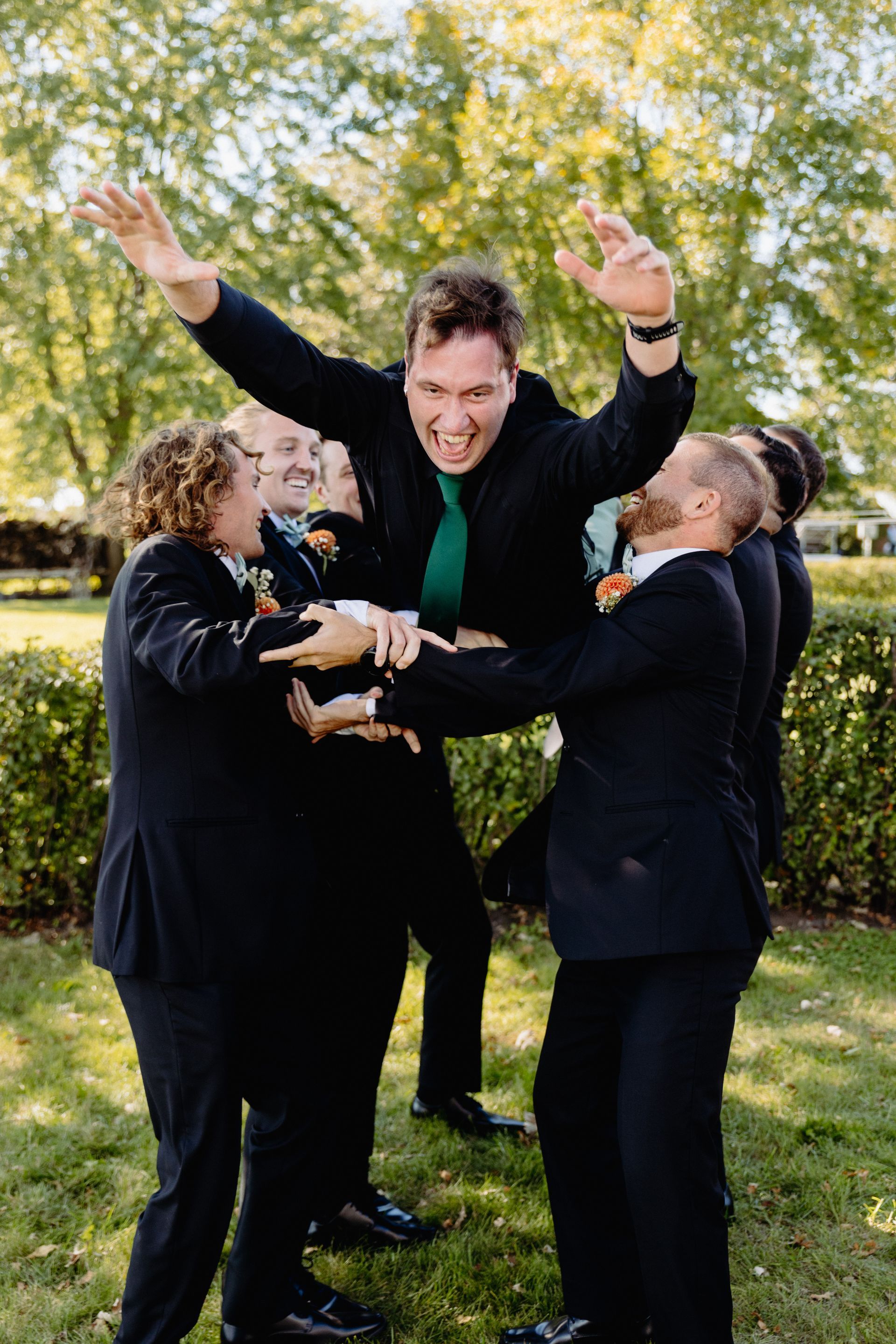 Groomsmen lifting the groom celebrating outdoors. He wears a green tie and raises his arms, smiling.