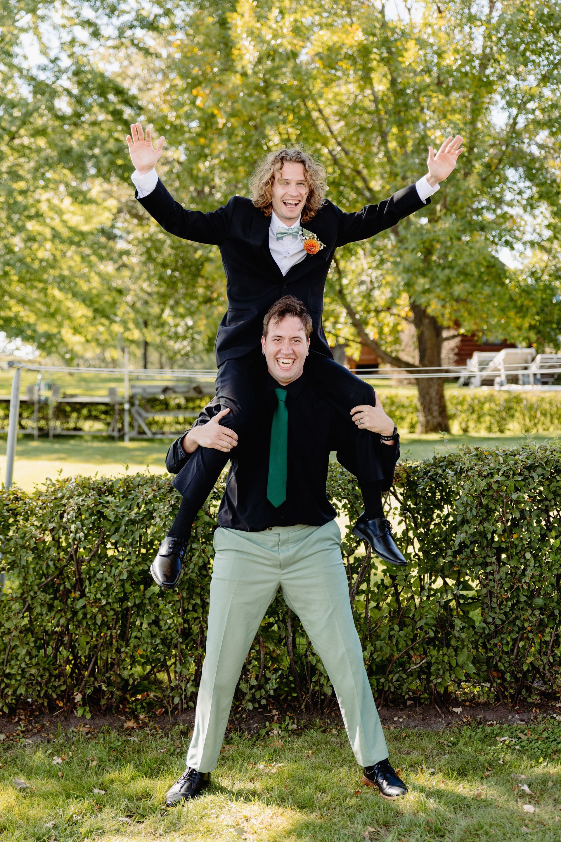 Two men joyfully, one on the other's shoulders; outdoors. Man on shoulders has arms raised, other smiles. Green foliage.