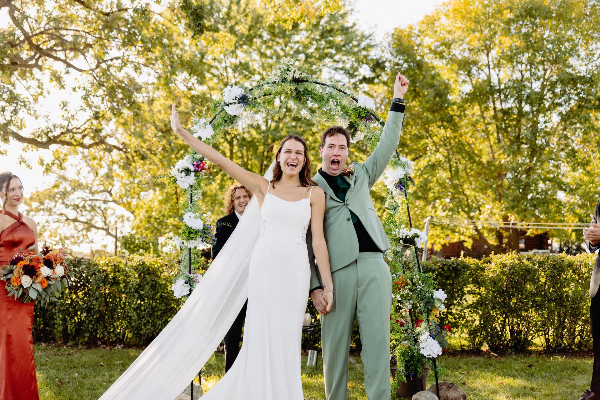Newlyweds celebrate; bride in white dress, groom in green suit, raise arms in joy. Outdoor ceremony.