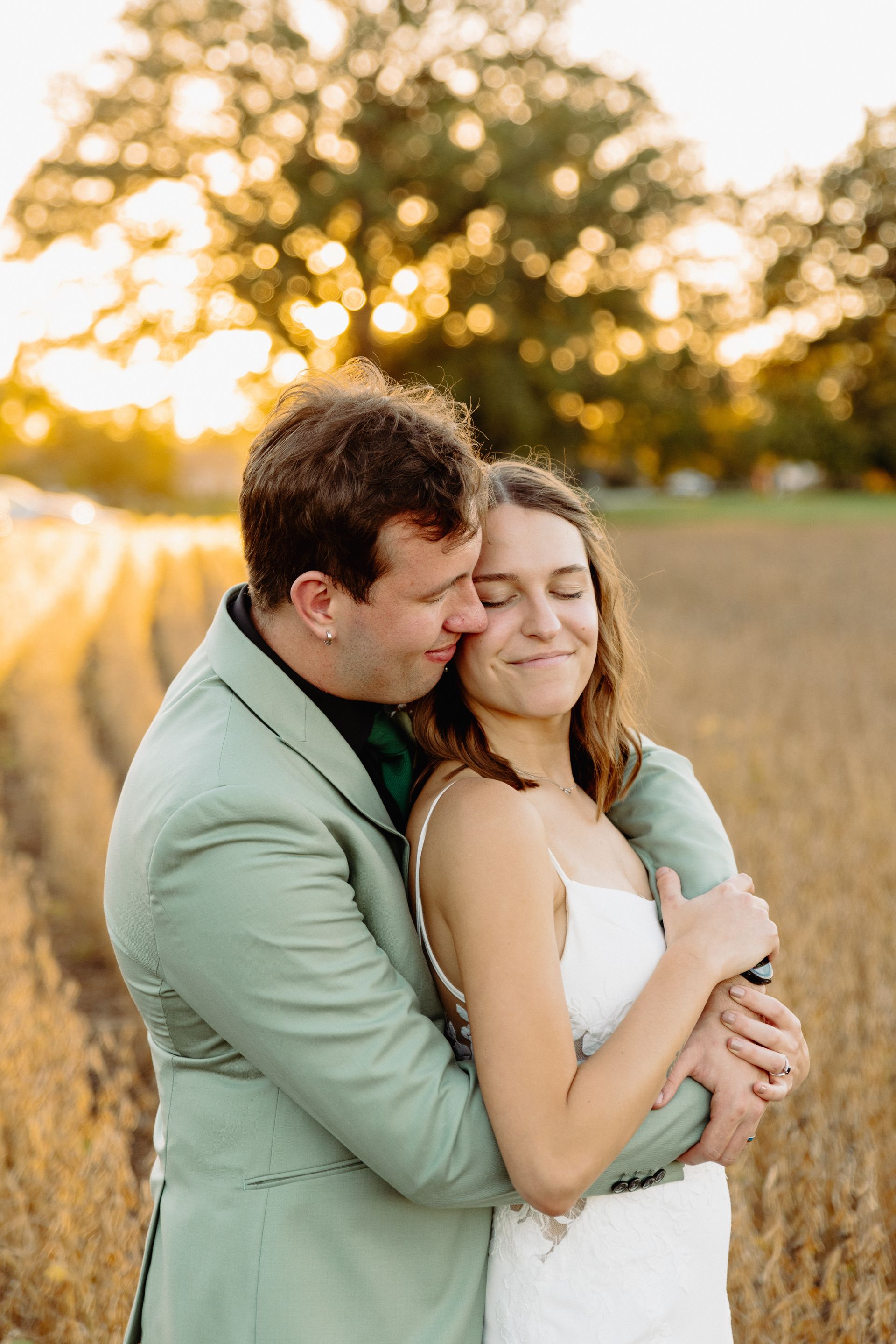 Couple embraces in a field. Man in green suit, woman in white dress, both smiling. Golden light.