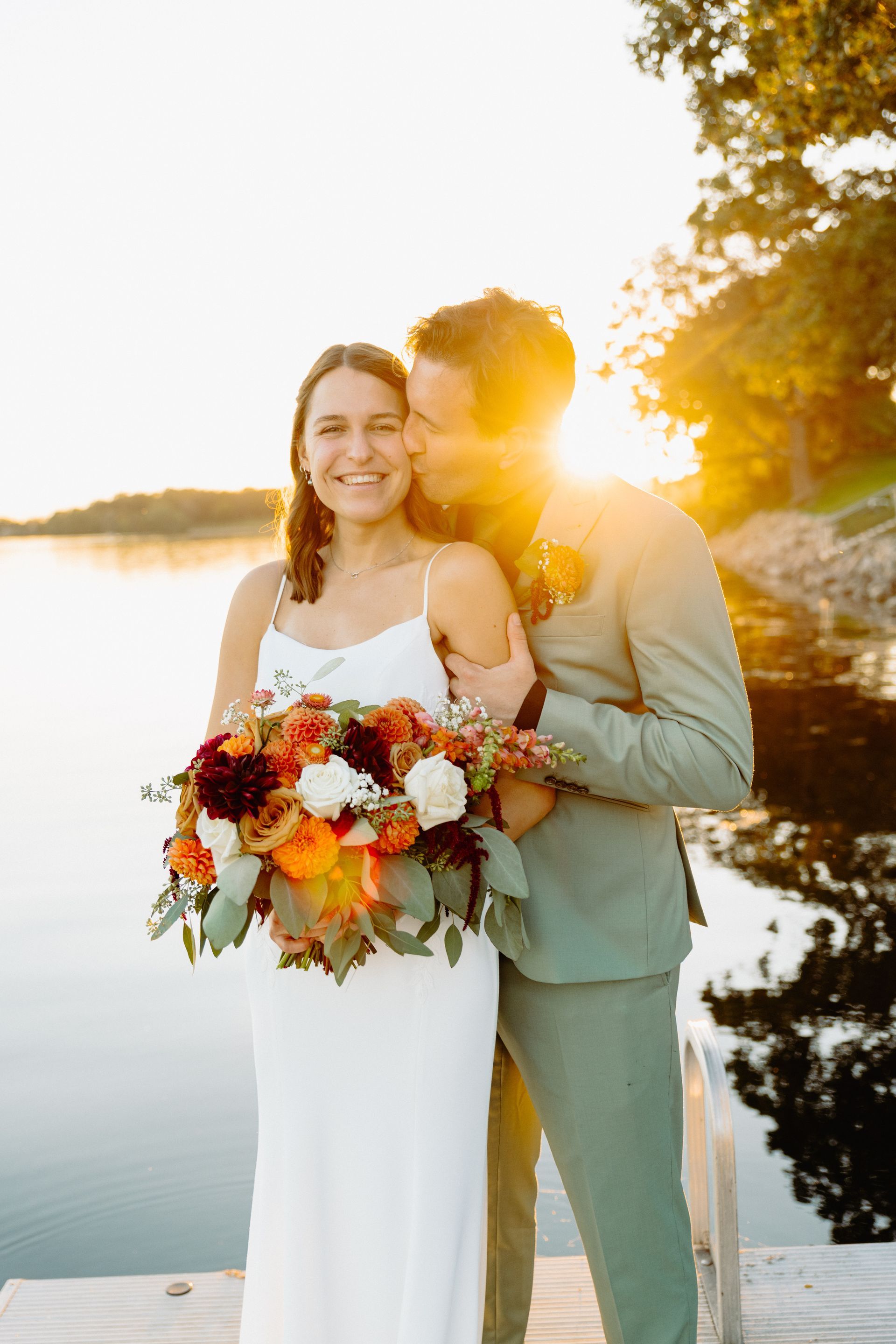 Bride and groom smile on a dock at sunset. Bride holds bouquet. Groom kisses her cheek, wearing a green suit.