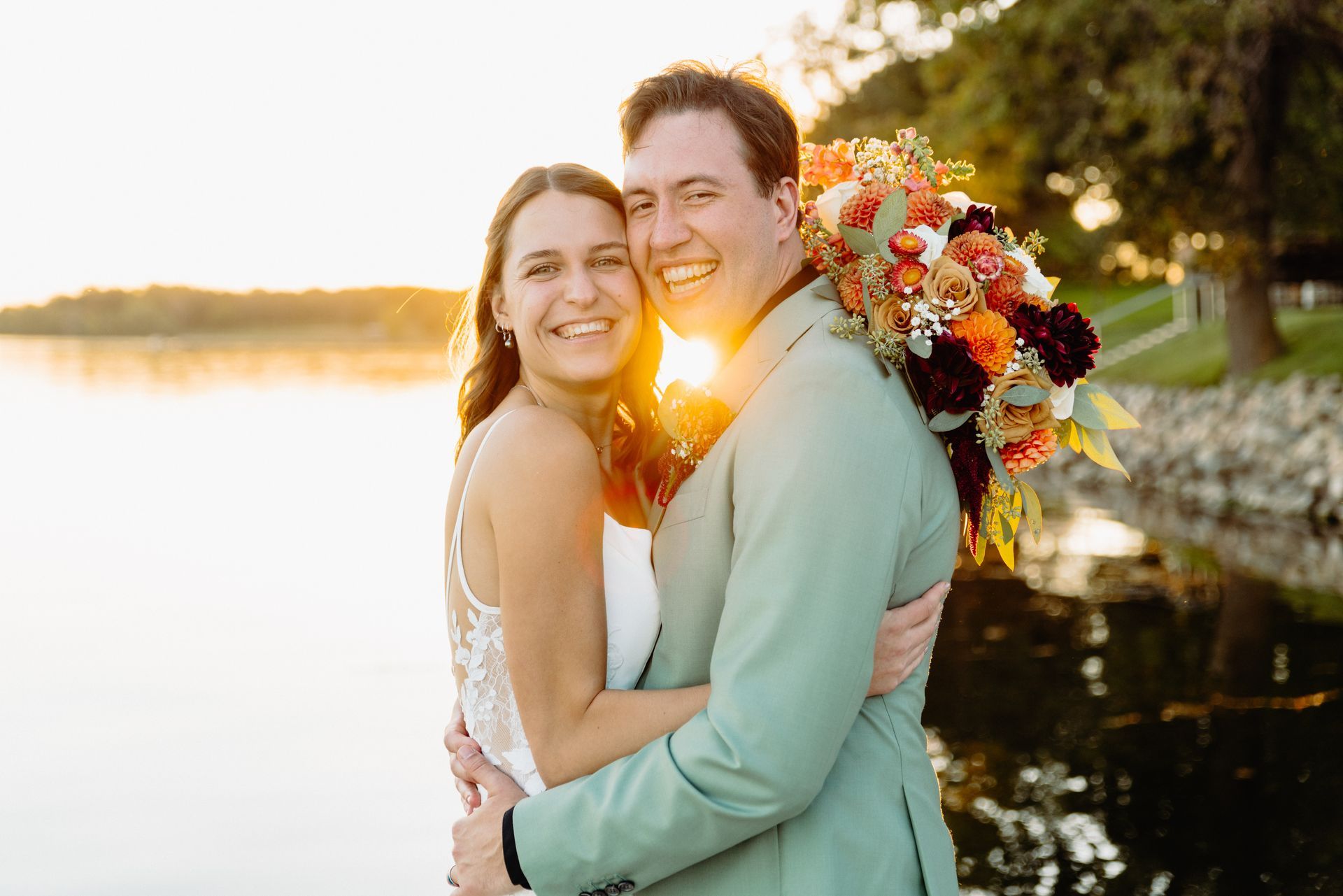 Newlyweds embrace, smiling near water at sunset. The bride holds a colorful bouquet.