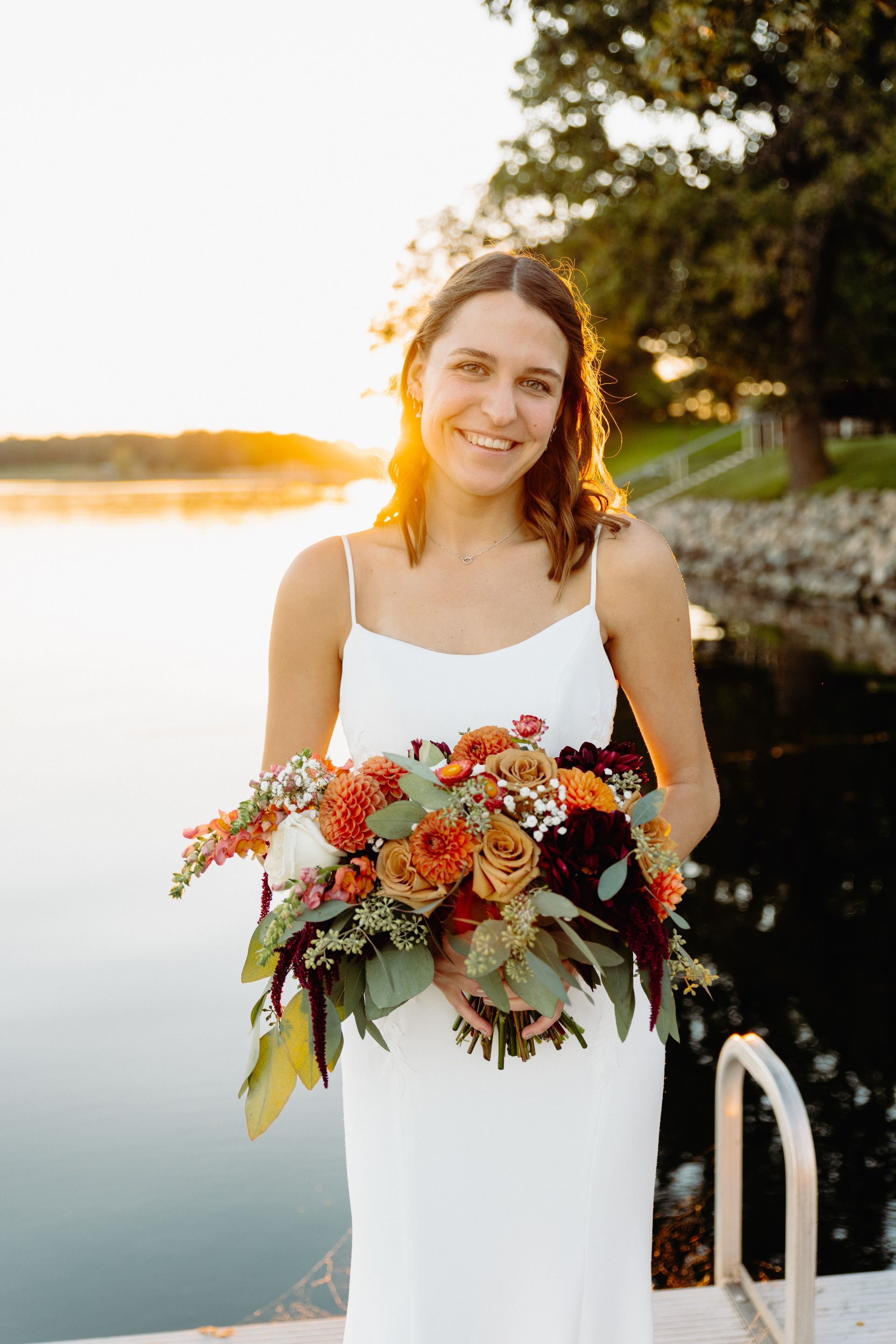 Woman in white dress holding colorful bouquet, smiling at sunset by lake.