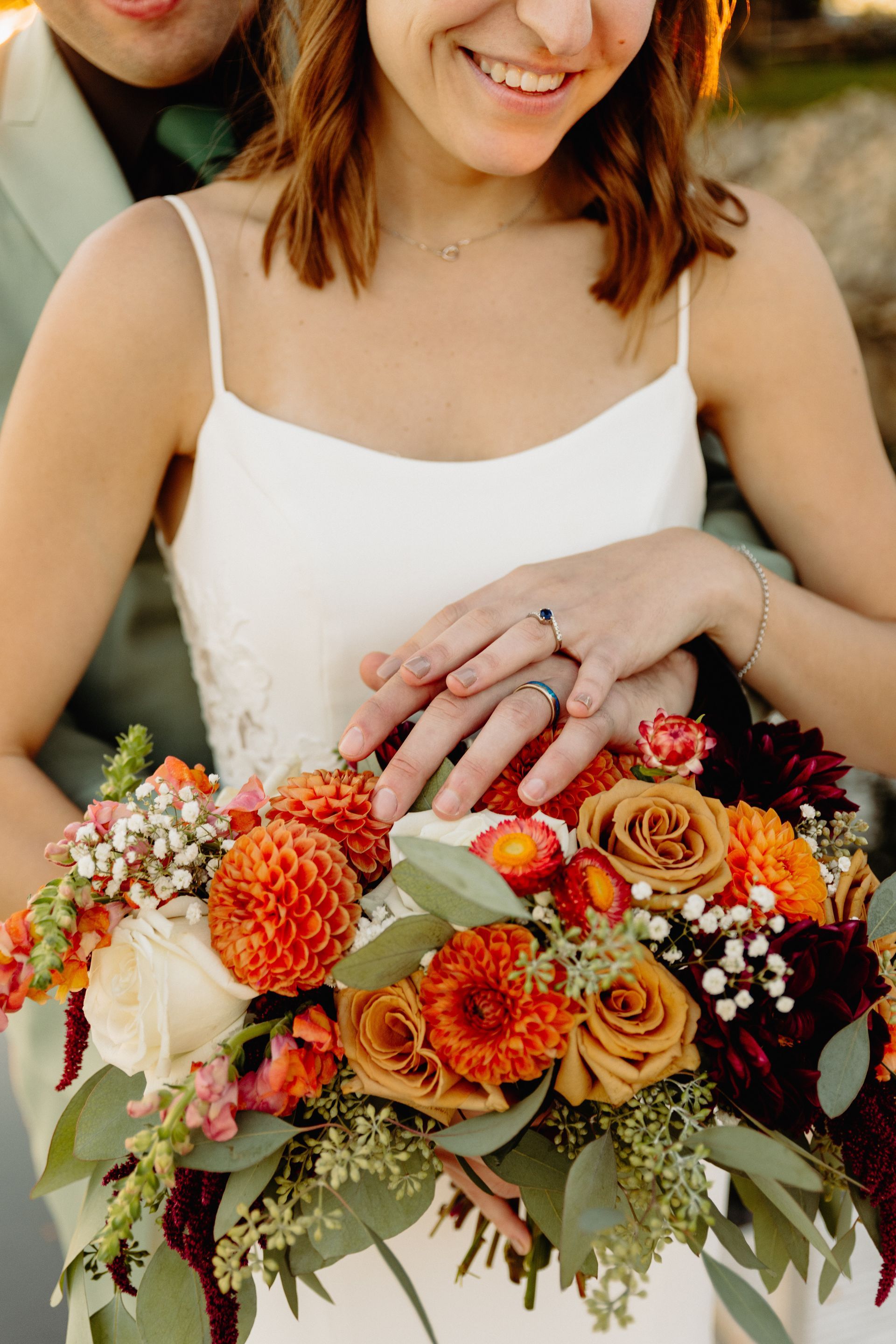 Couple with hands together over colorful wedding bouquet, smiling, outdoor setting.