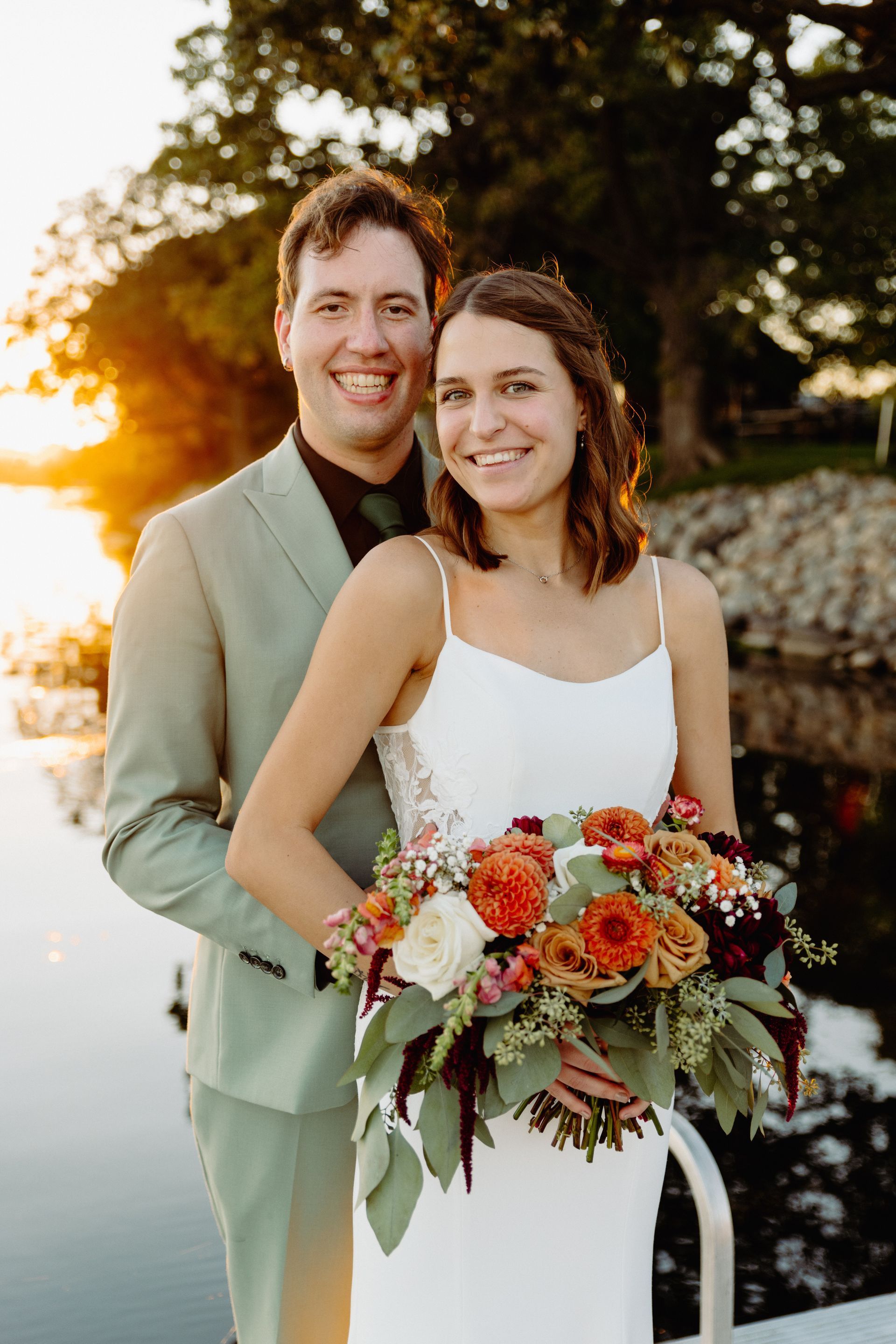 Newlywed couple smiling; woman in white dress holds bouquet; man in light green suit, sunset background.