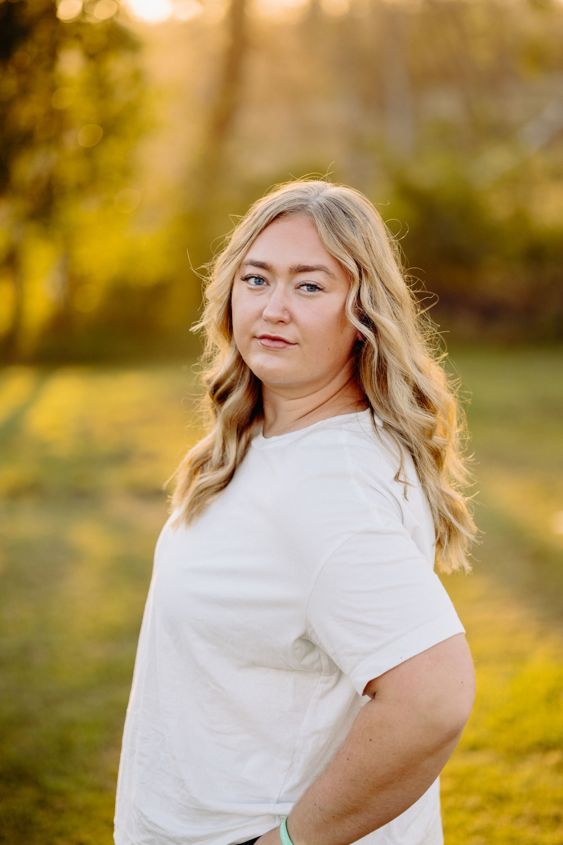 Woman with blonde wavy hair, wearing a white shirt, standing outside in front of blurred trees.