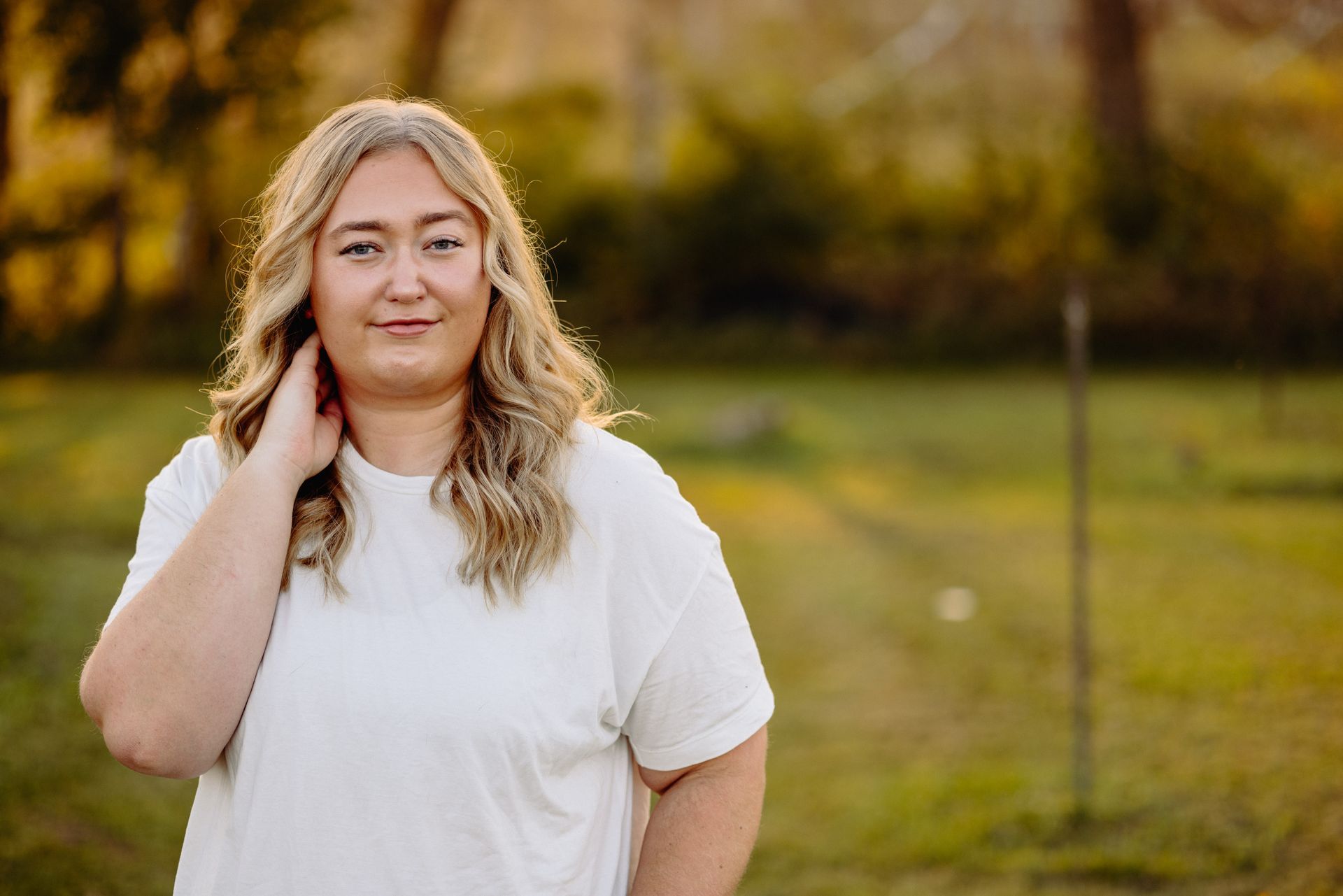 Woman with blonde hair, wearing a white t-shirt, poses outdoors with hand near her neck, smiling in front of a blurred green background.