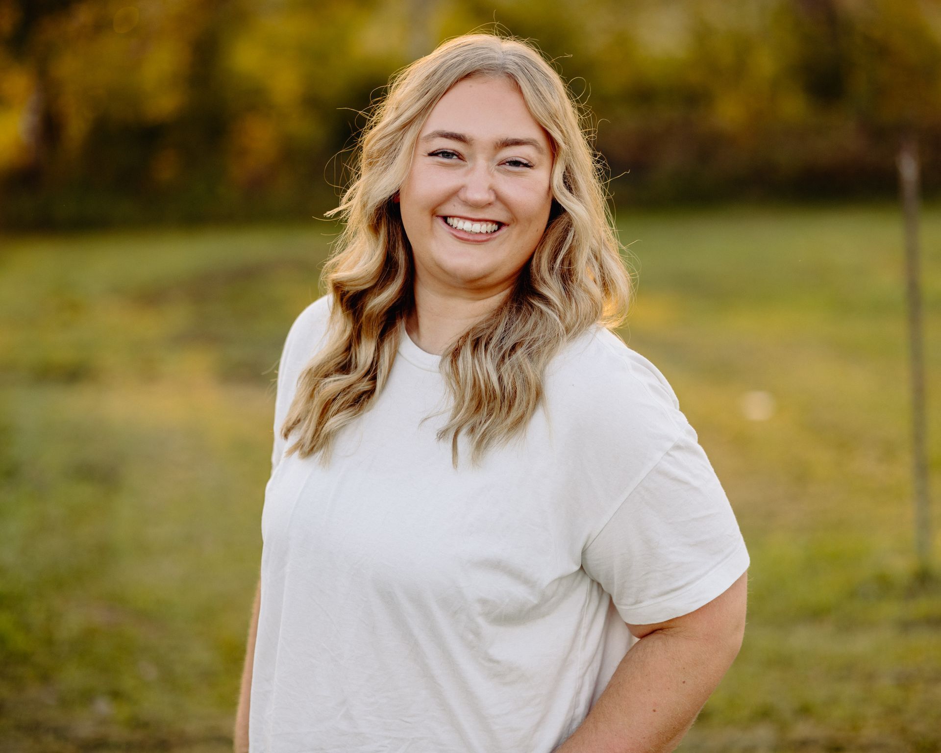 Smiling woman with blonde hair in a field, wearing a white t-shirt, arm at her side.