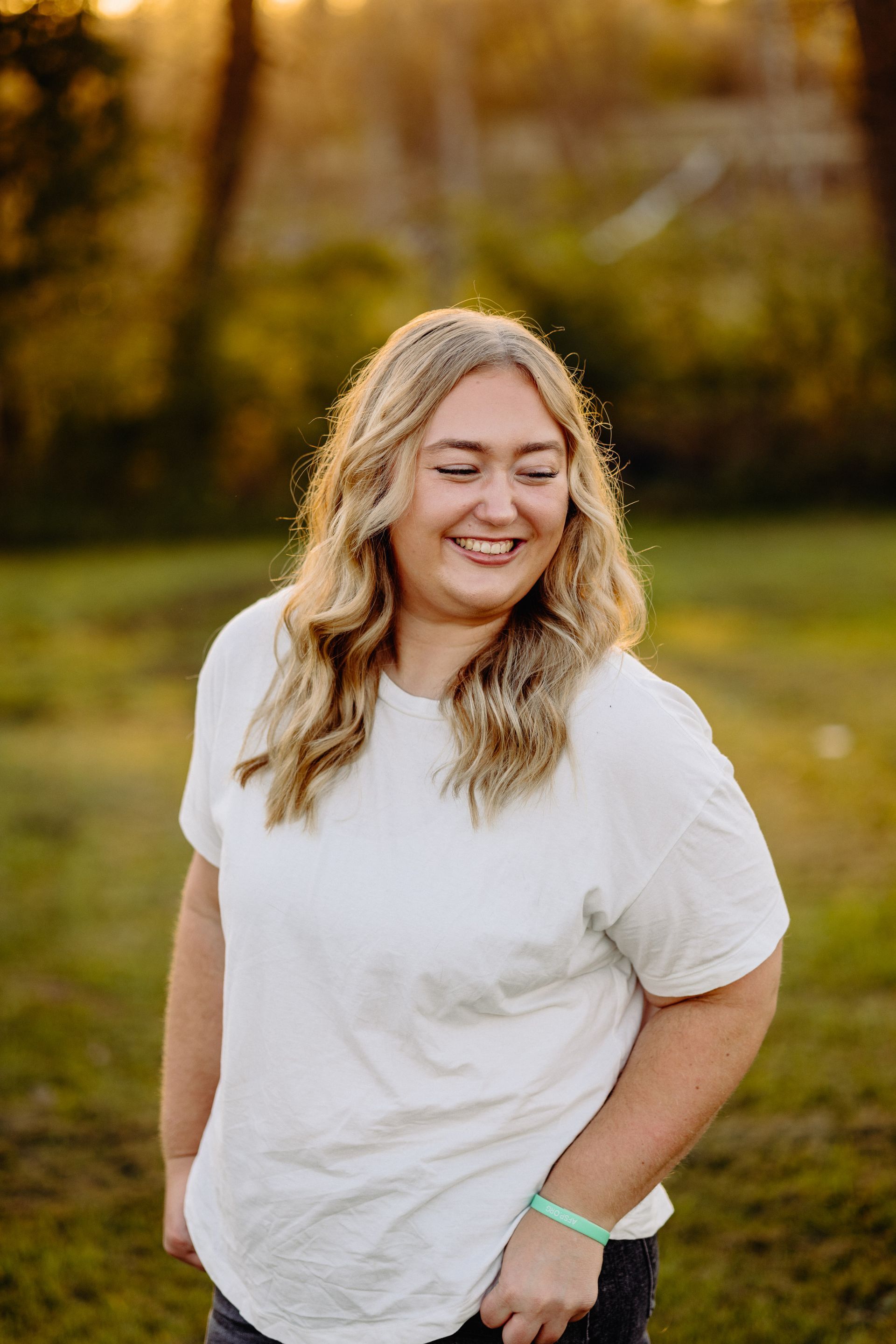 Blonde woman smiling outdoors, wearing white shirt, hand in pocket, sunlight in background.