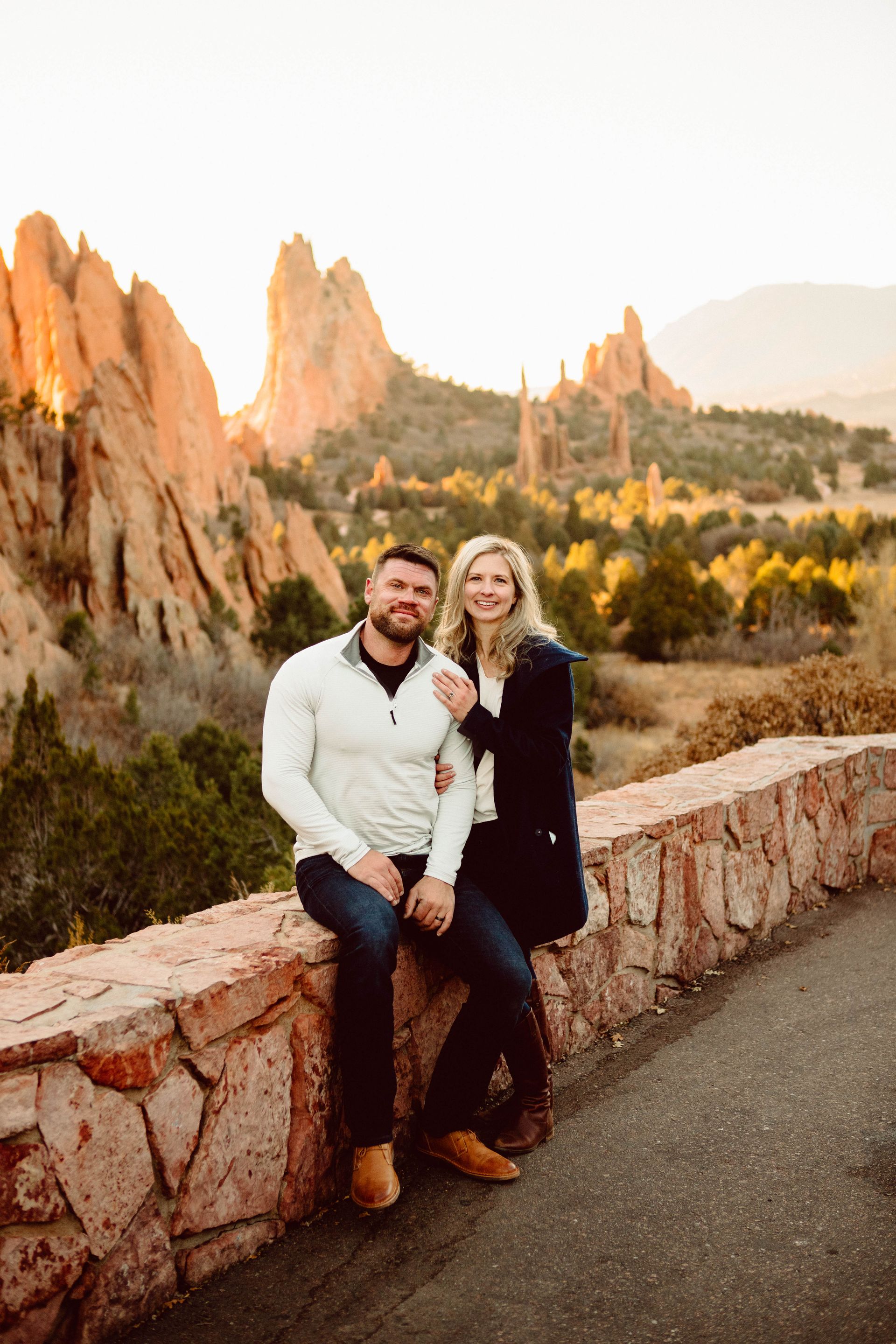 Couple sitting on a stone wall, smiling, with red rock formations in the background.