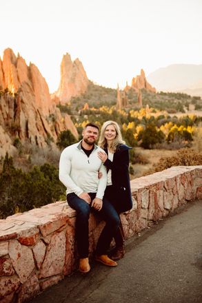 Couple smiling, posing on a stone wall; red rock formations in the background at sunset.
