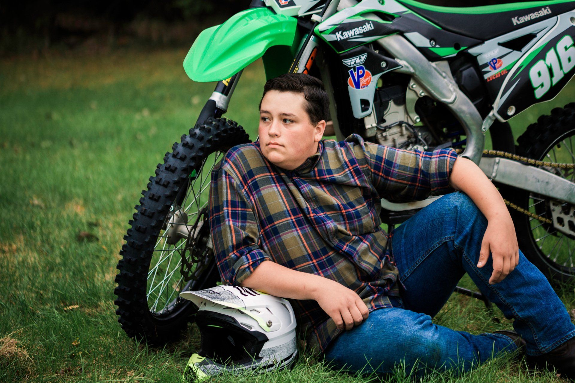 Young person in flannel shirt and jeans leans against a green and black dirt bike on grass.