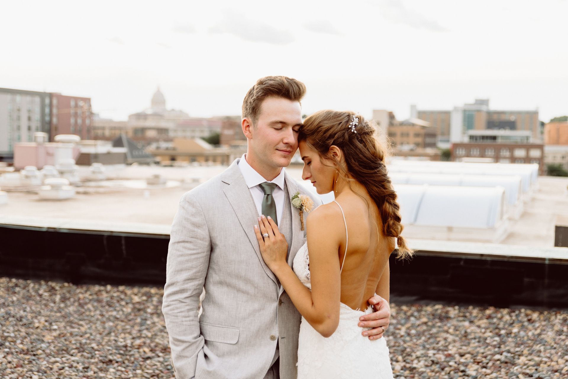 Newlyweds embrace on a rooftop; the bride in a backless dress, the groom in a striped suit, city buildings in background.