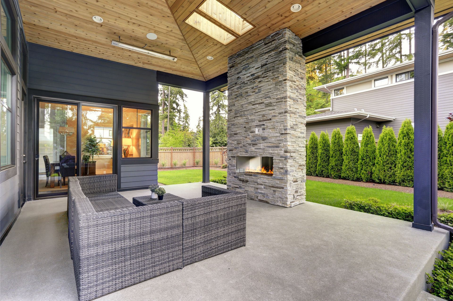 Covered patio with wicker seating, stone fireplace, and view of a green backyard