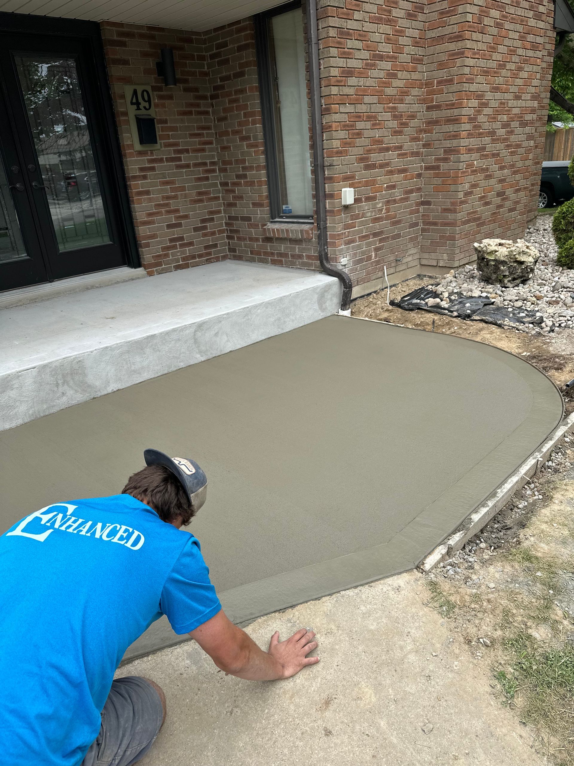 A man in a blue shirt is working on a concrete walkway in front of a brick house.
