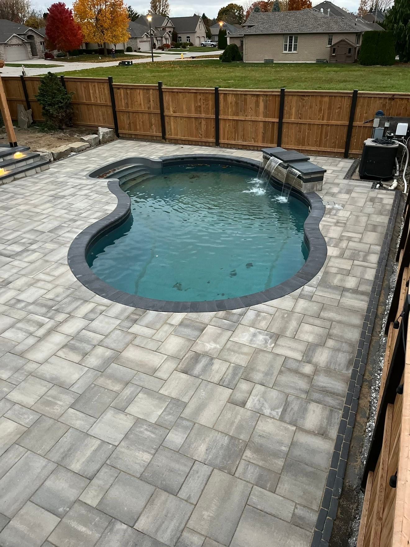 A large swimming pool is surrounded by a brick patio and a wooden fence.