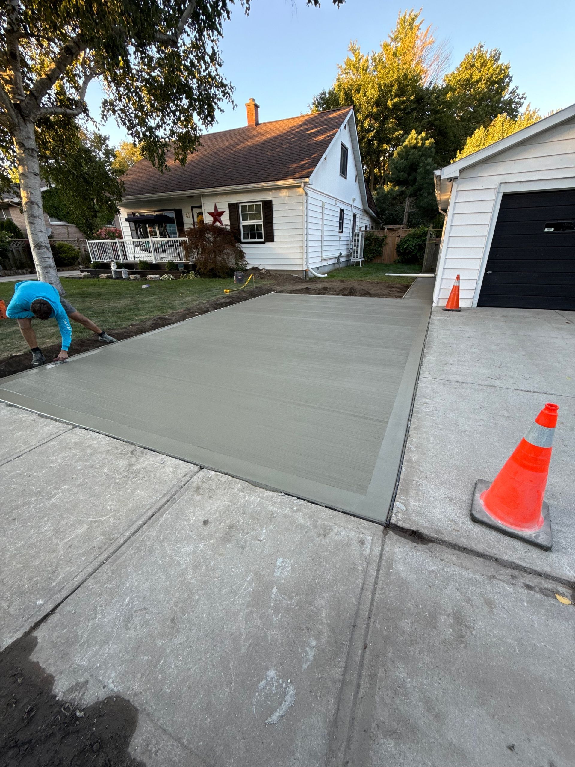 Freshly poured concrete driveway beside a sidewalk, orange safety cones, worker finishing edges.