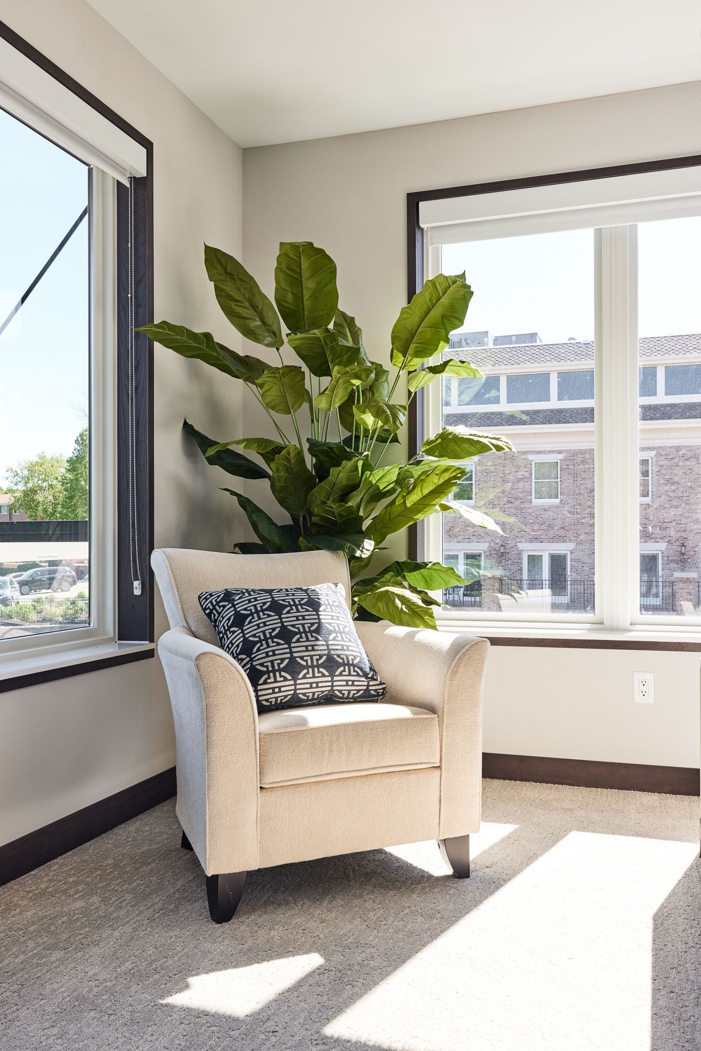 Cozy corner with a cream armchair, decorative pillow, and tall green plant near windows.