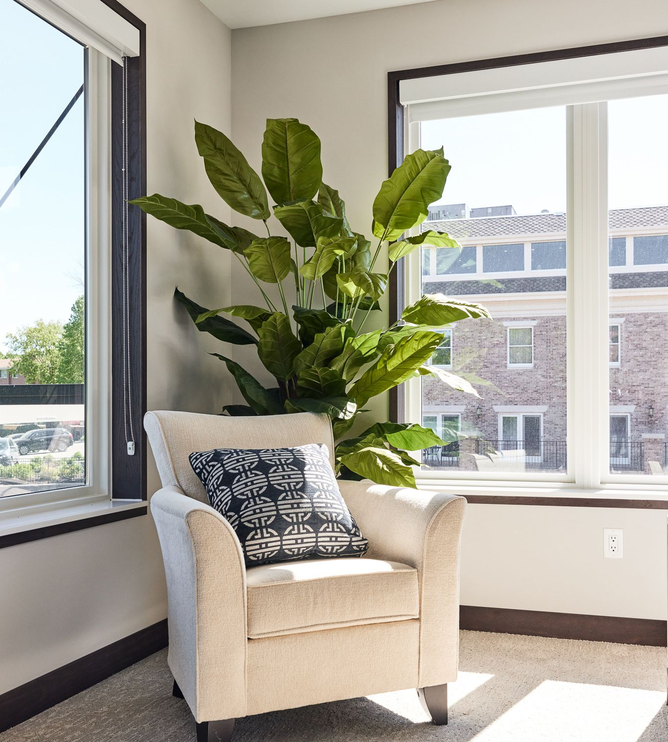 Beige armchair with a decorative pillow, next to large green plant and windows.
