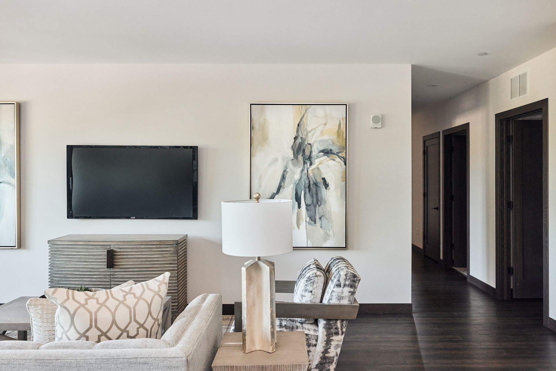 Living room with mounted TV, art, lamp, and neutral-toned furniture. Hallway with dark doors and floor visible.