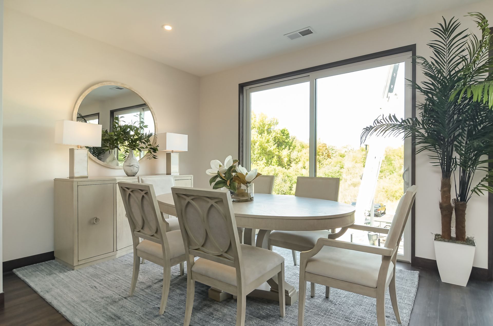 Dining room with a round table, chairs, a buffet, and a large window with trees outside.