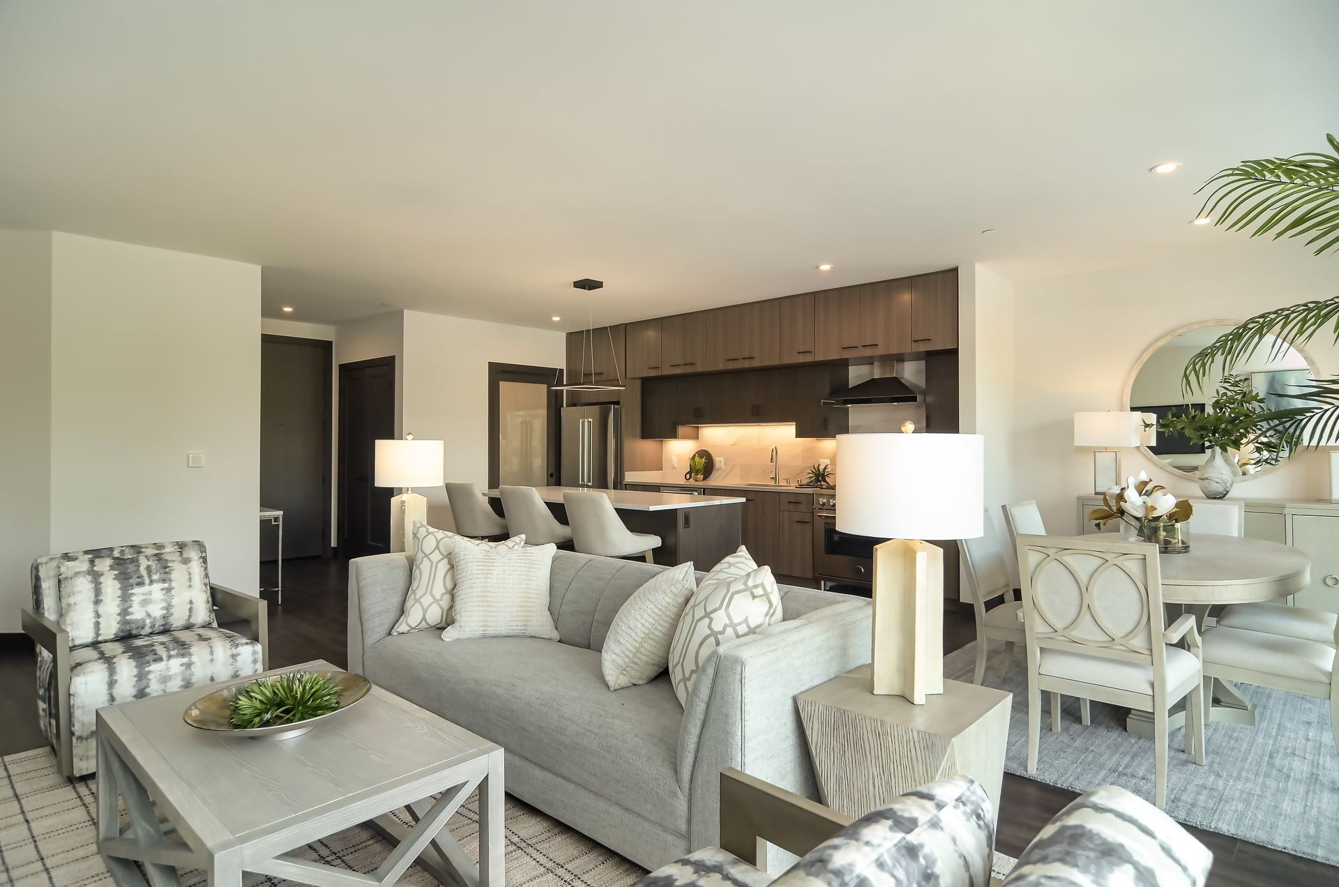 Open-concept living space with gray sofa, kitchen, dining area, and neutral decor.