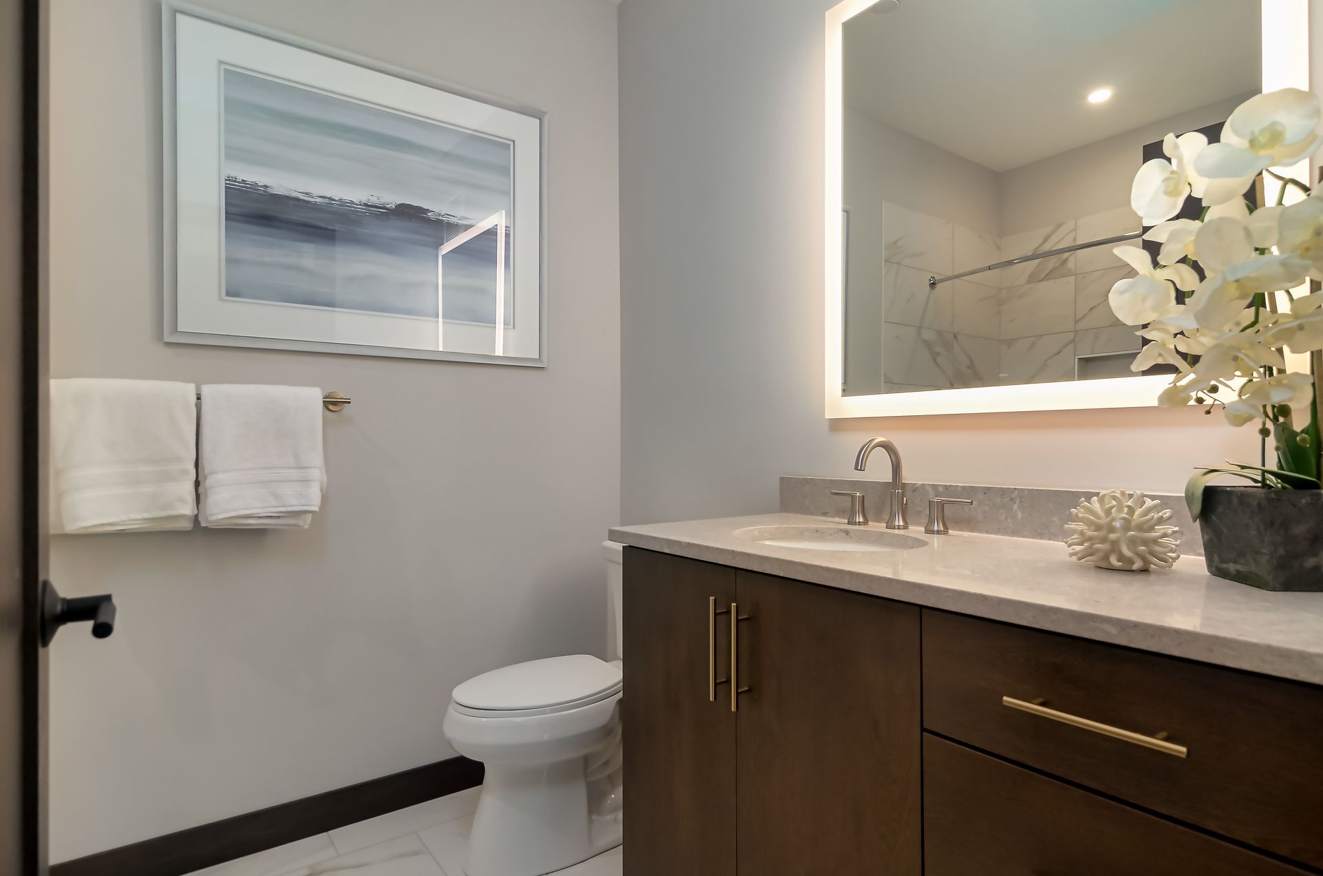 Bathroom with a large illuminated mirror, white countertop, dark wood vanity, toilet, and decorative orchid.
