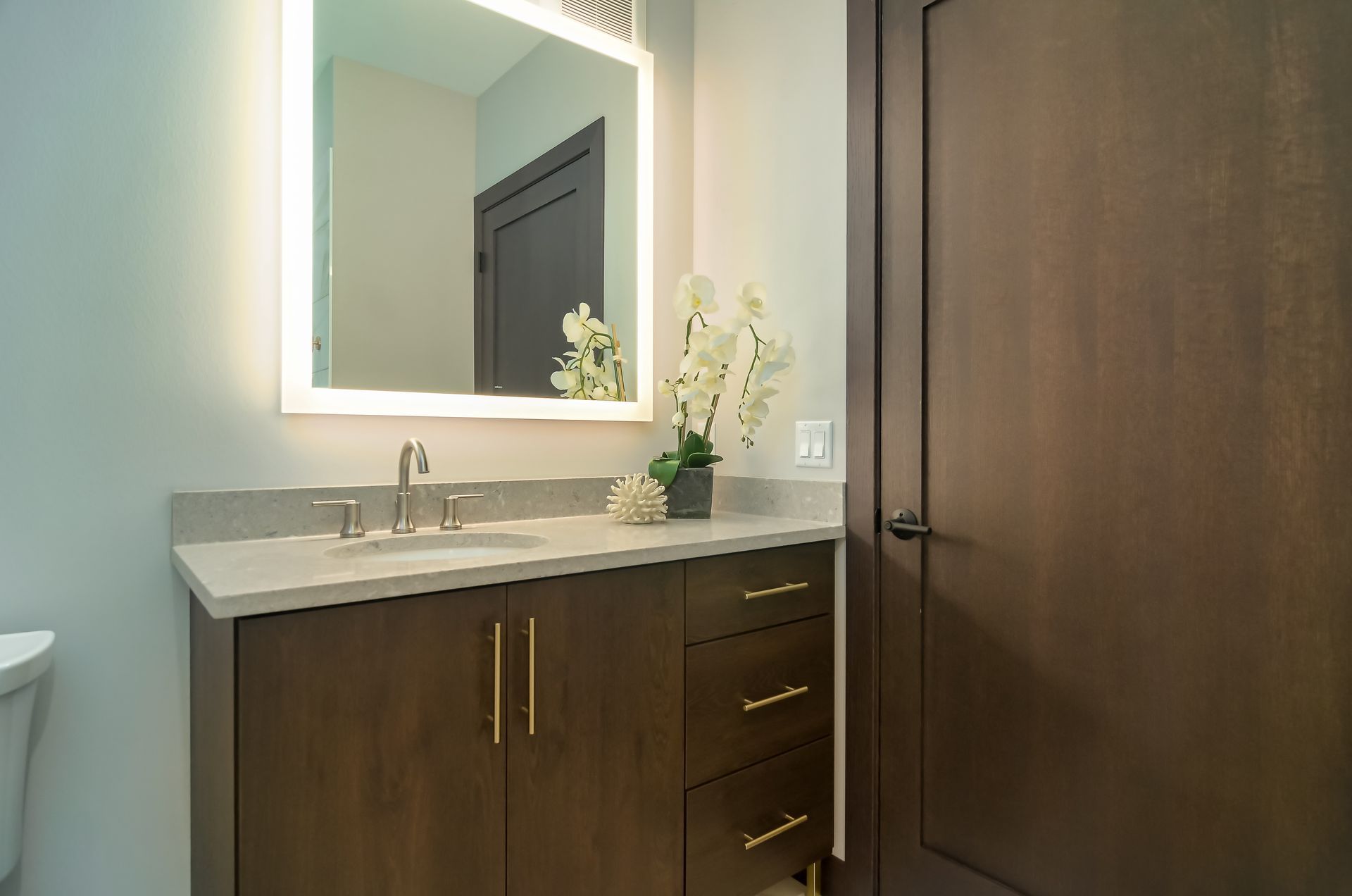 Bathroom vanity with a lighted mirror, brown cabinets, and a white countertop.