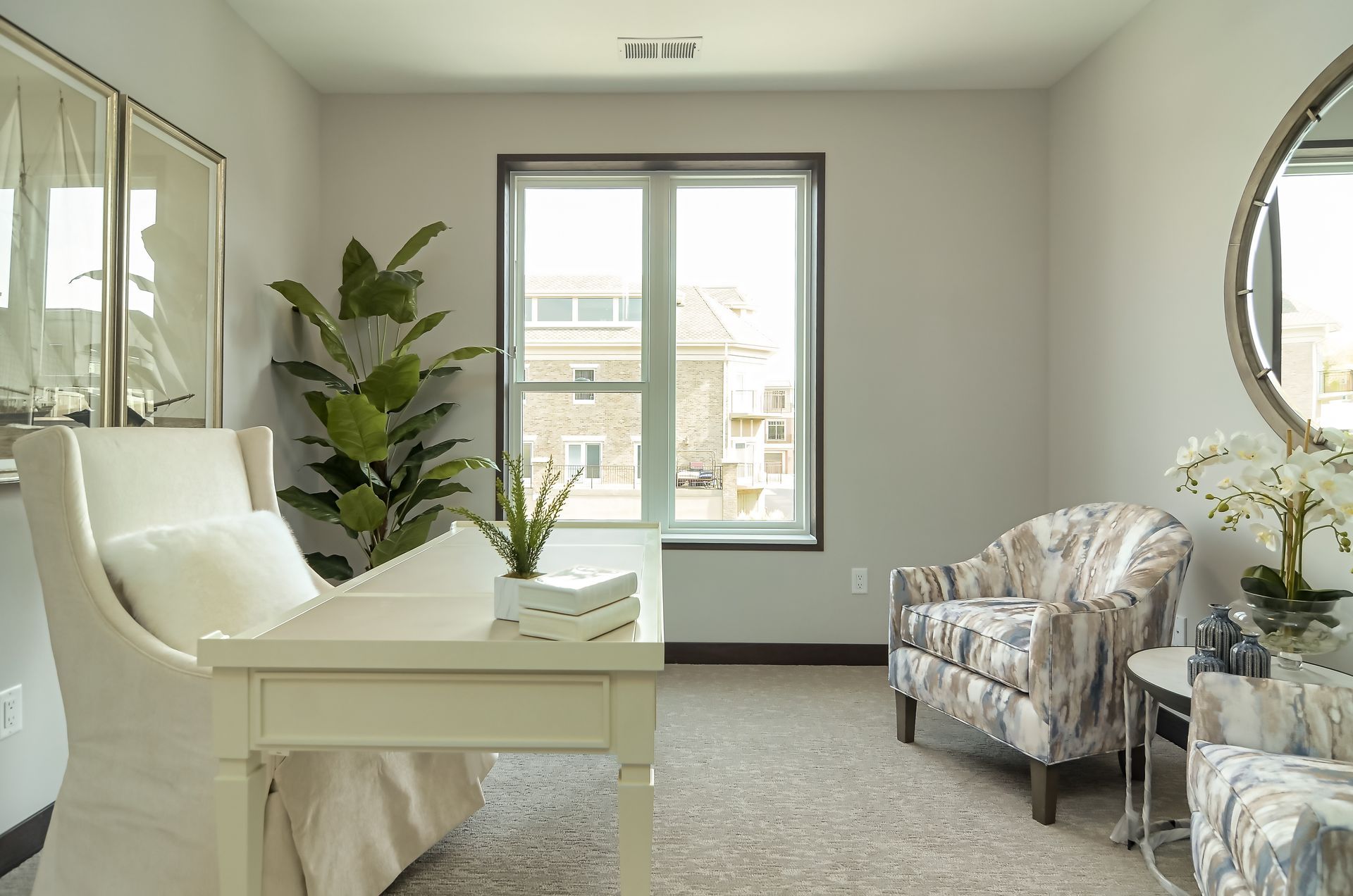 Light-filled home office with white desk, patterned armchairs, tall plant, and framed art. Window offers an outside view.
